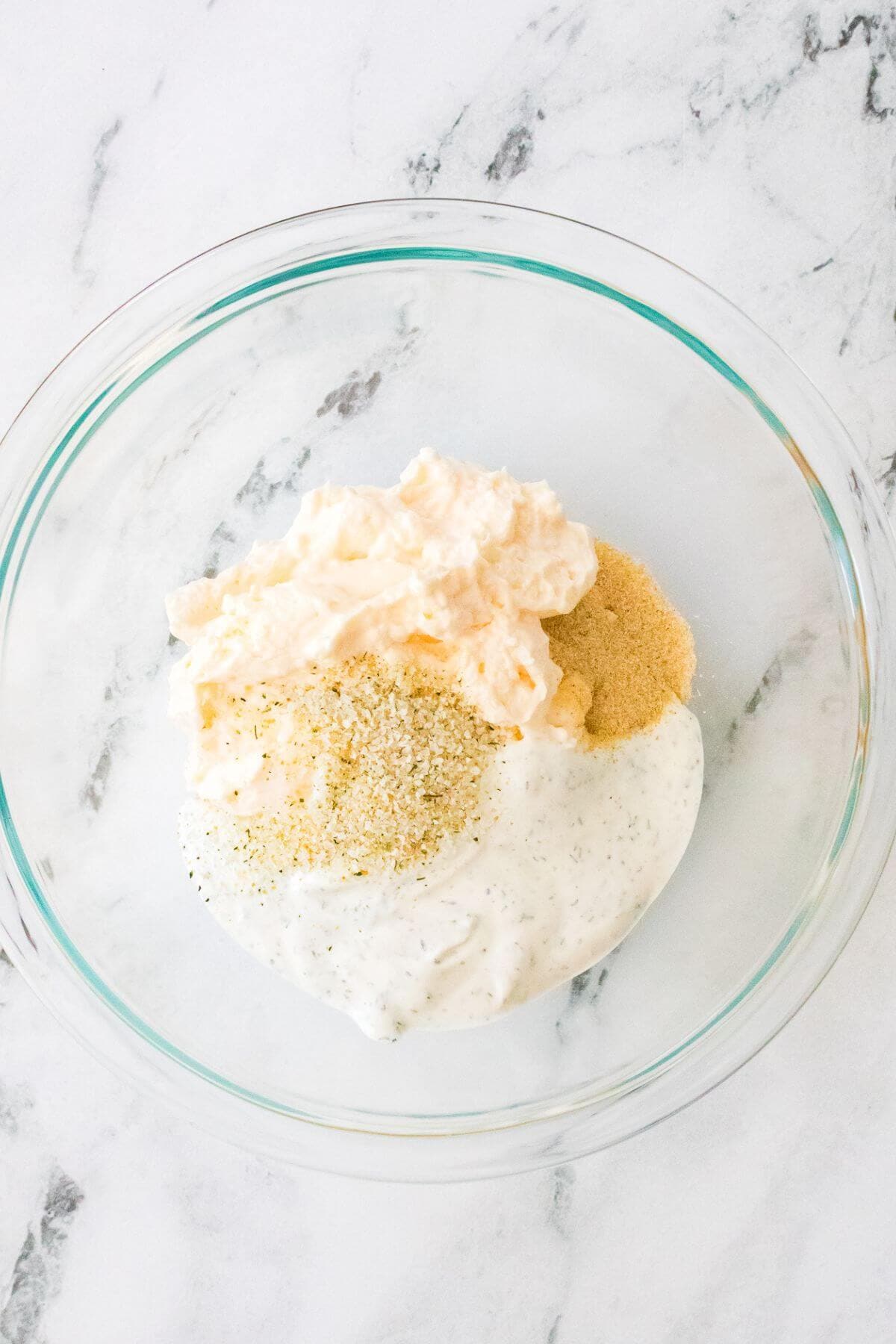 A glass bowl containing mayonnaise, grated cheese, yogurt, and breadcrumbs on a marble surface.