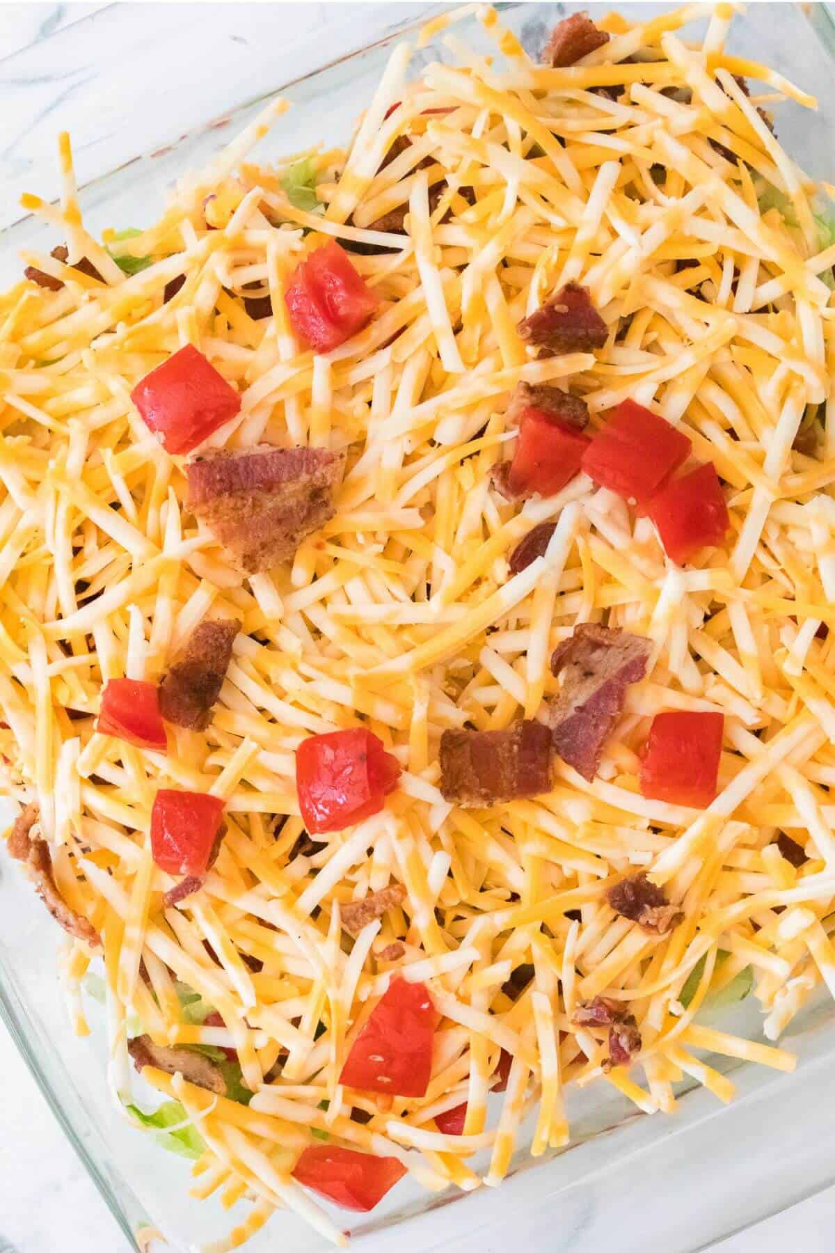 A glass dish filled with shredded cheese, diced tomatoes, and bacon bits.