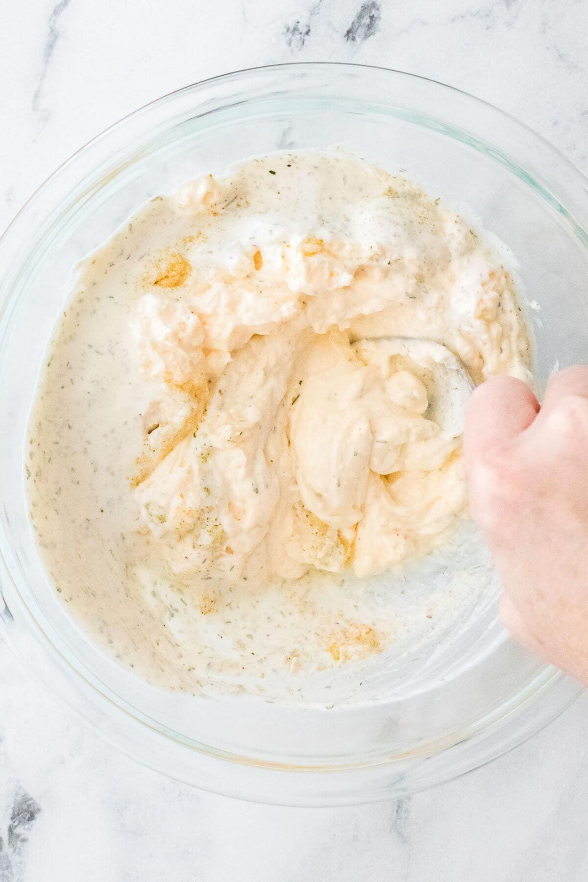 A hand mixing creamy white and orange dip in a glass bowl on a marble counter.