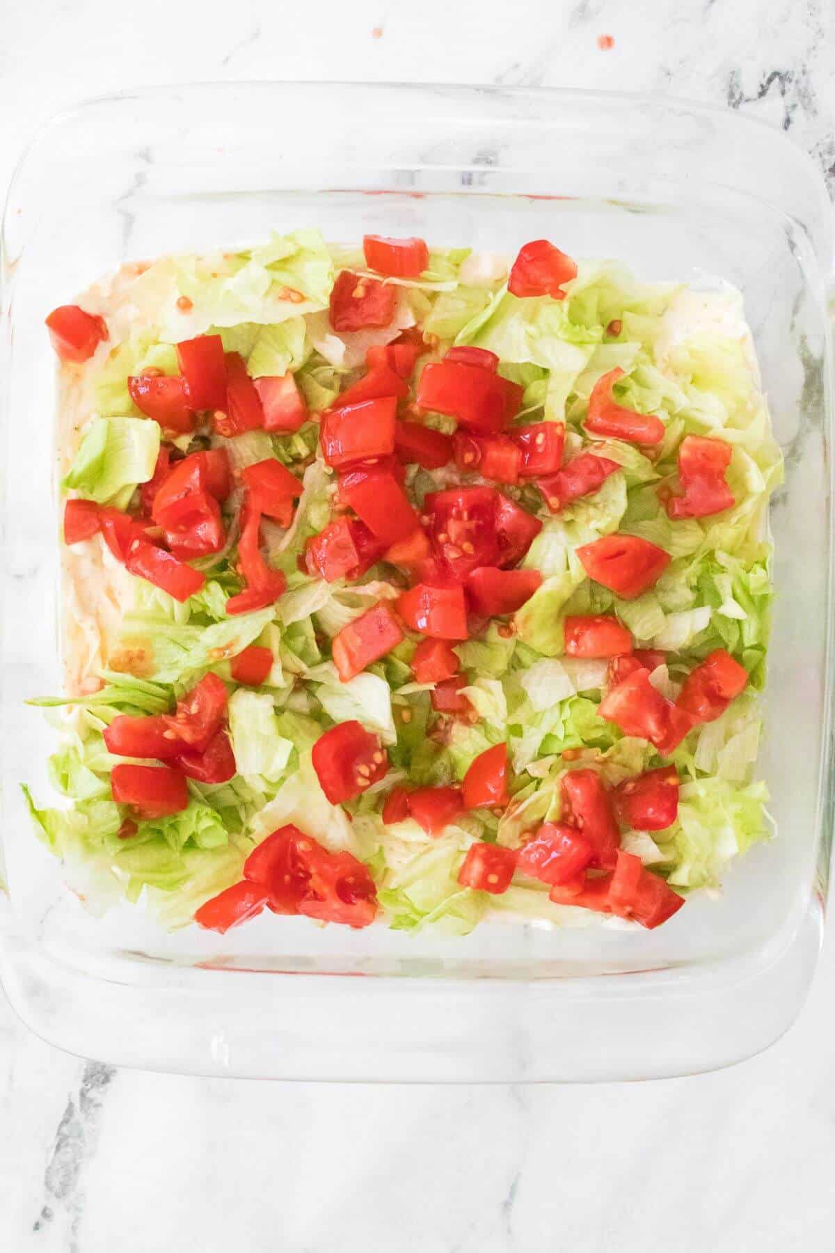 Glass dish with layered dip topped with chopped lettuce and diced tomatoes on a marble surface.
