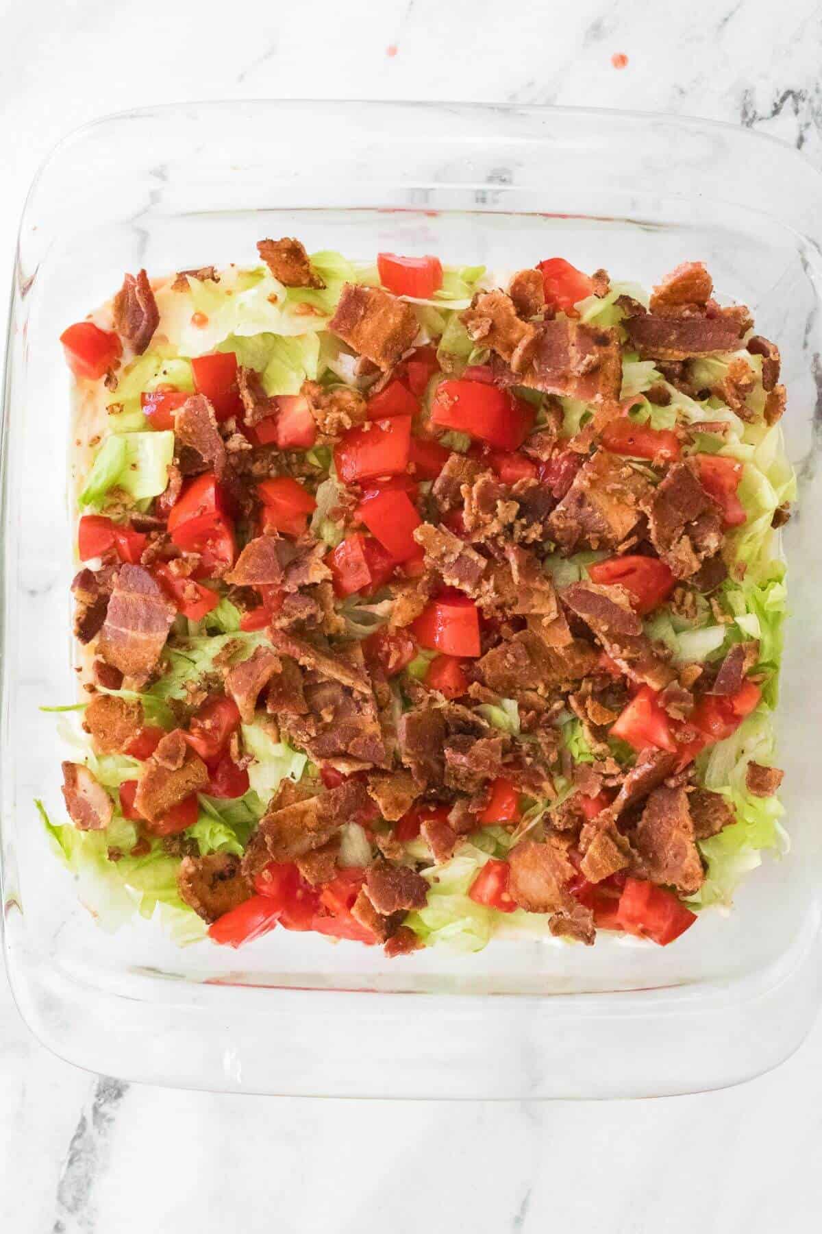 A square glass dish holds a layered salad topped with lettuce, chopped tomatoes, and crumbled bacon.