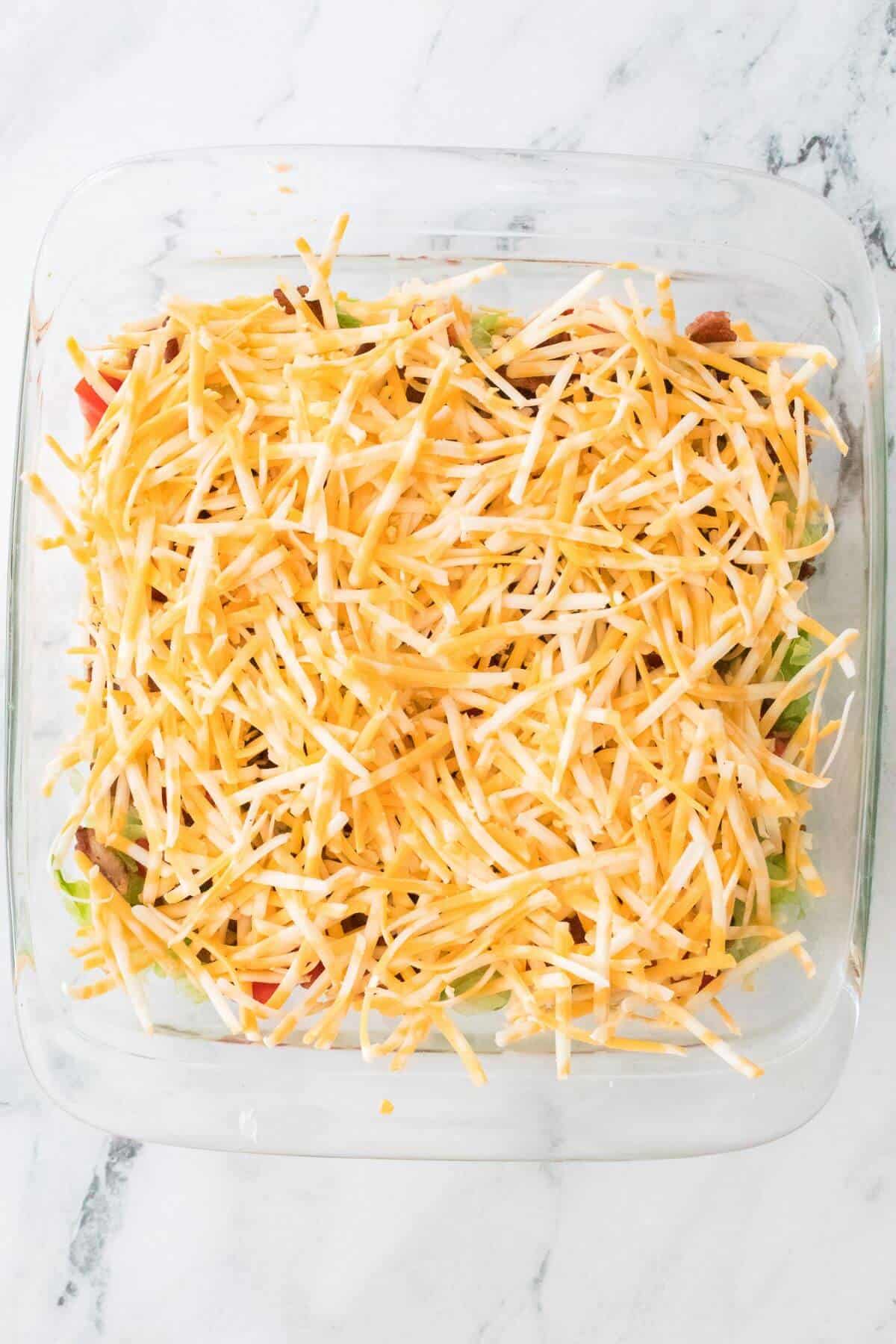 A clear glass baking dish filled with shredded cheese layered on top of sliced vegetables on a marble countertop.