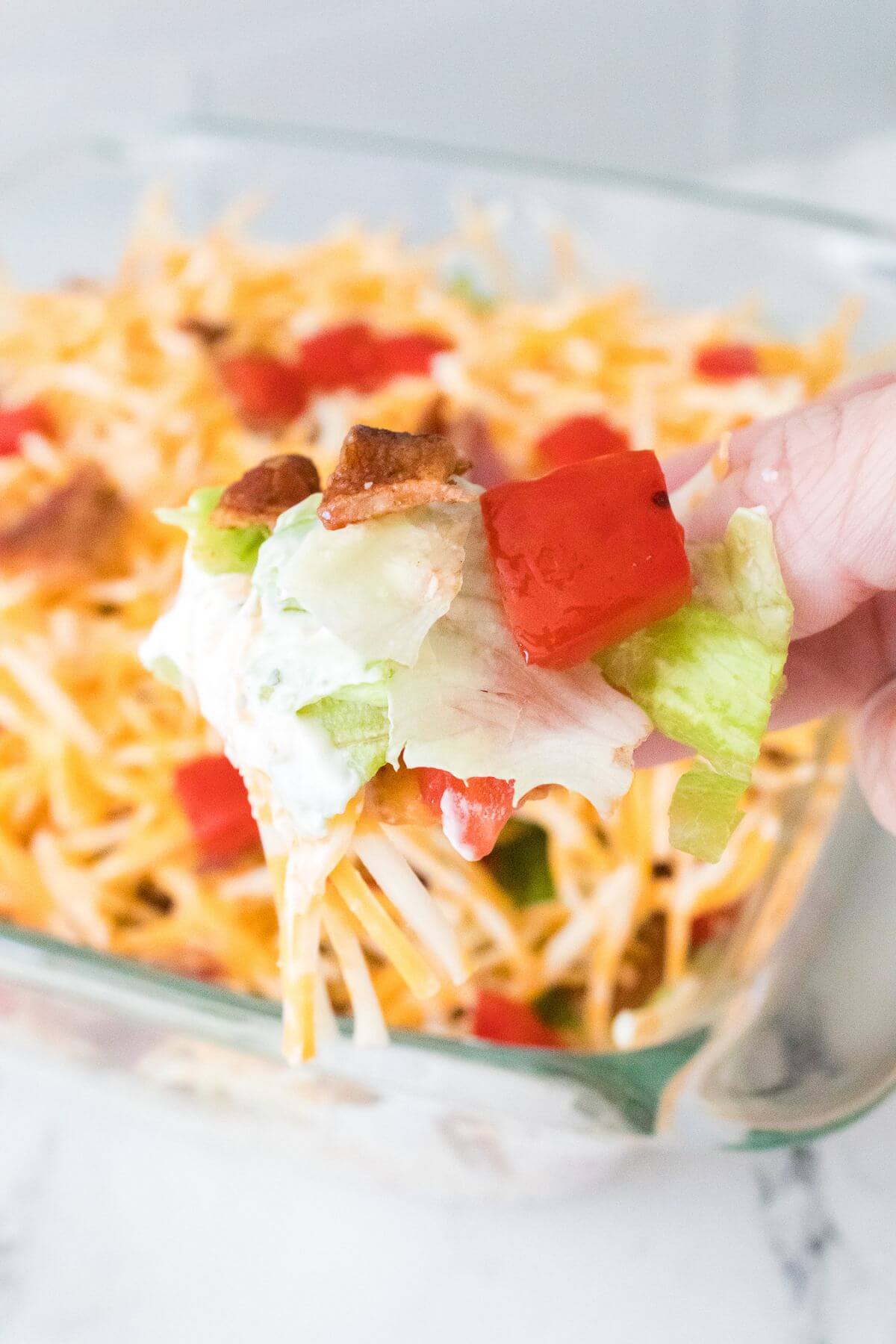 A close-up of a hand holding a lettuce scoop topped with shredded cheese, diced tomatoes, and bacon, above a full casserole dish of similar salad.