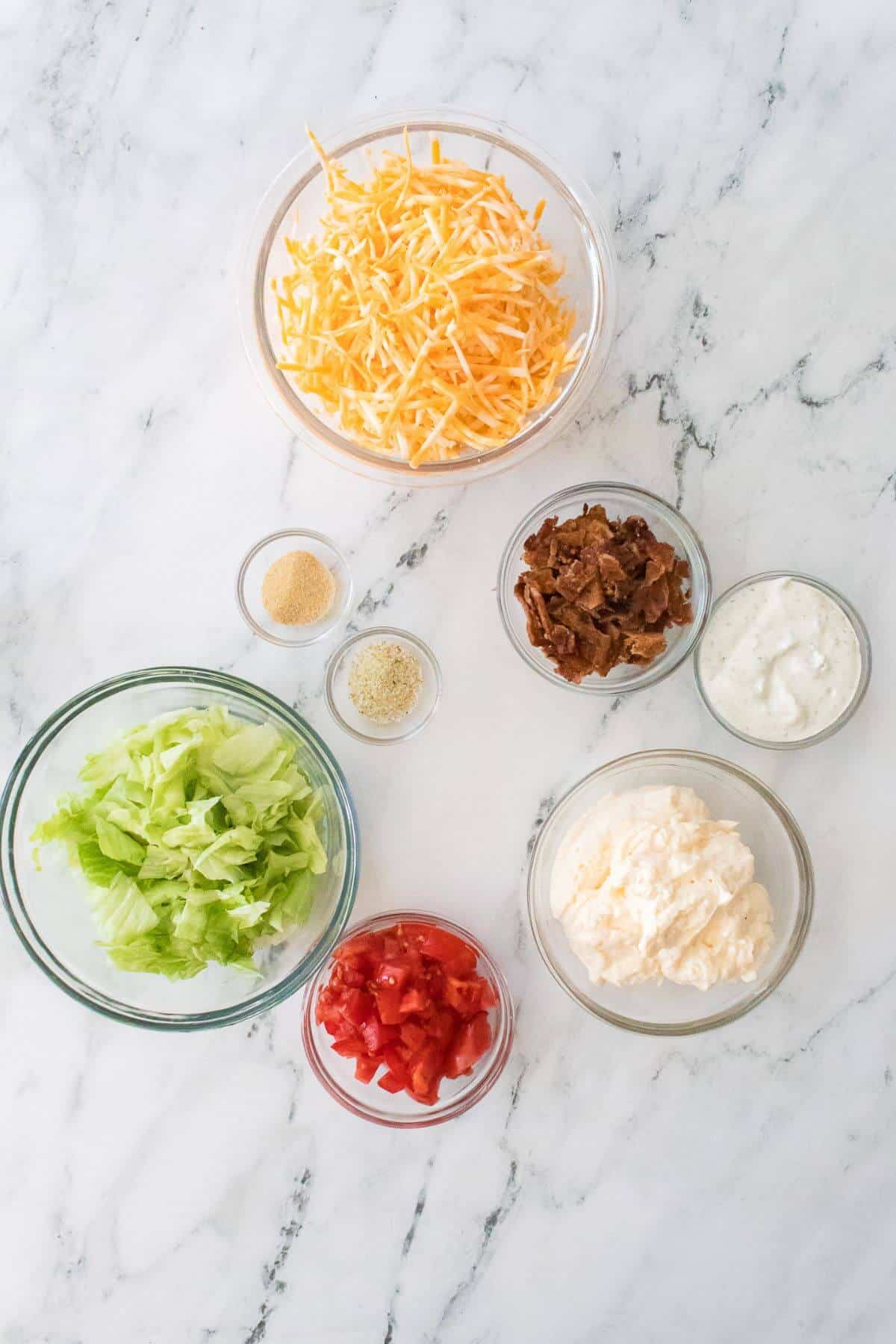 Ingredients for a salad including shredded cheese, lettuce, chopped tomatoes, bacon bits, mayonnaise, ranch dressing, onion powder, and garlic powder on a marble surface.