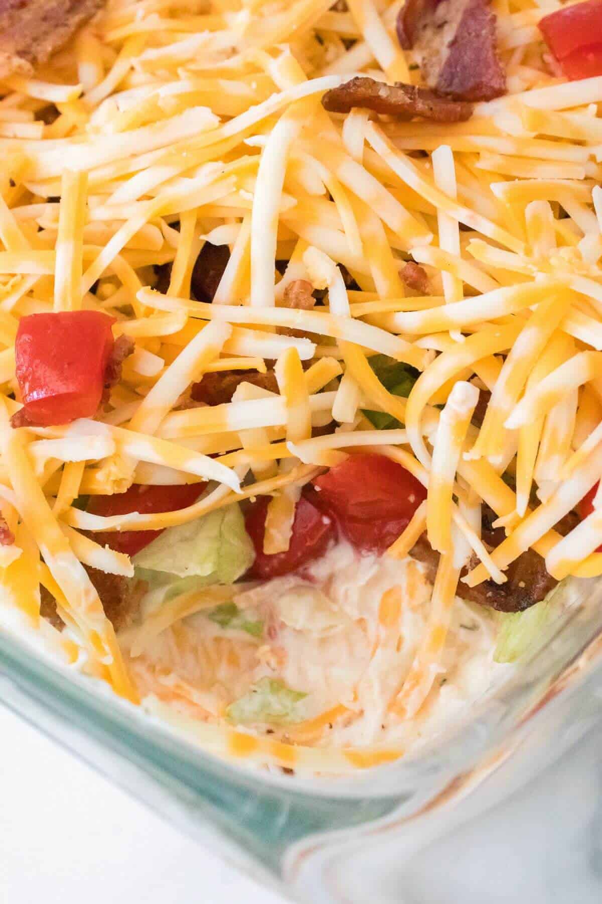 Close-up of a layered salad in a glass dish with shredded cheese, chopped tomatoes, and bacon on top.
