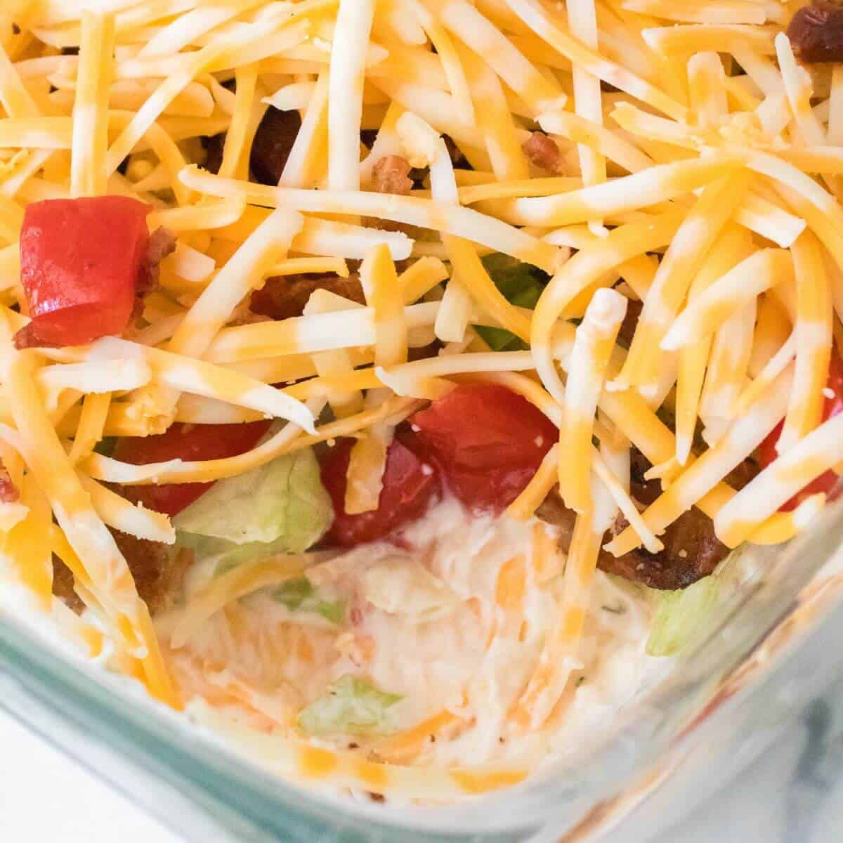 Layered salad with shredded cheese, chopped tomatoes, and visible greens in a glass dish.