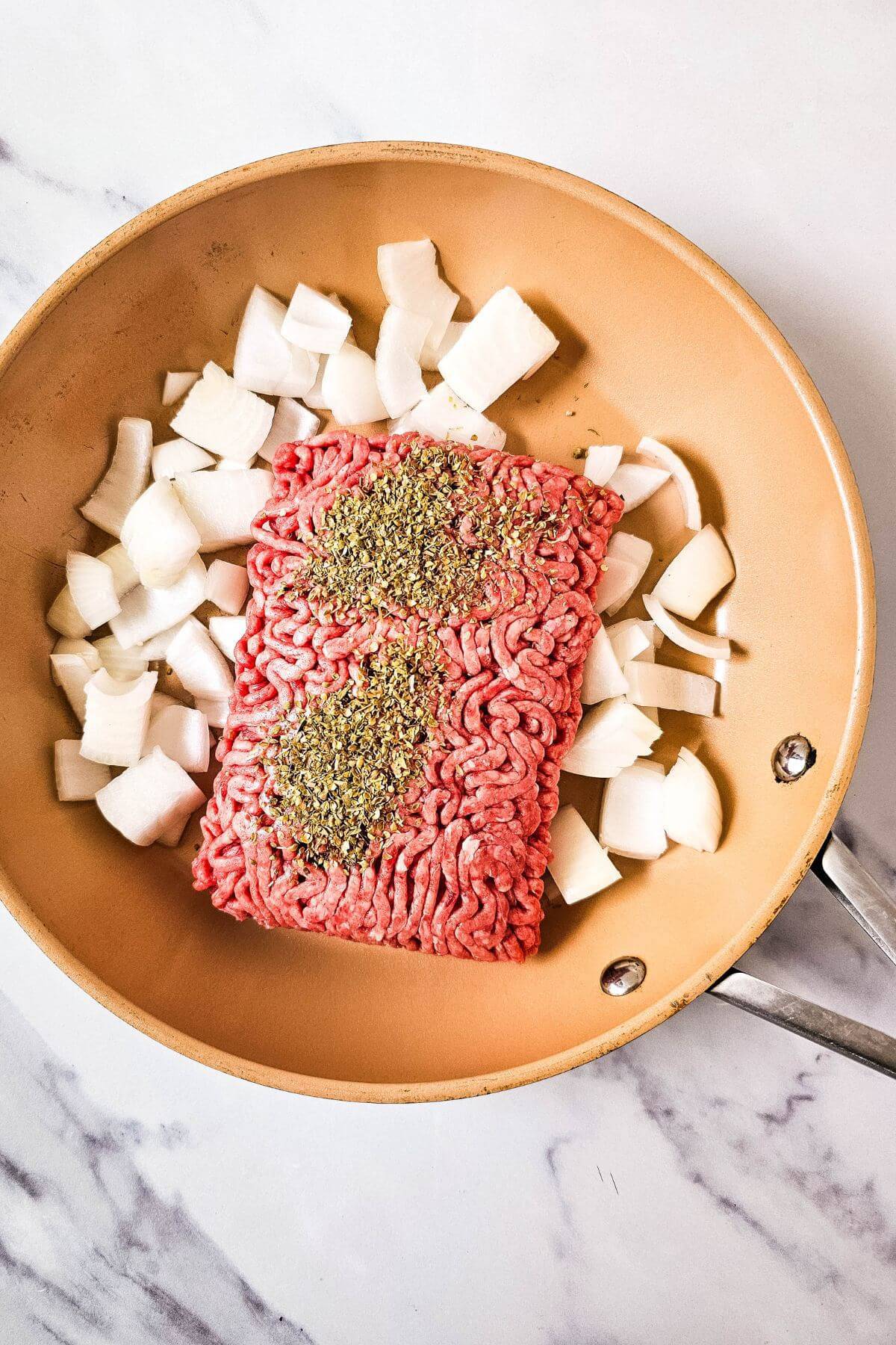 A skillet with raw ground beef topped with herbs, surrounded by chopped onions.