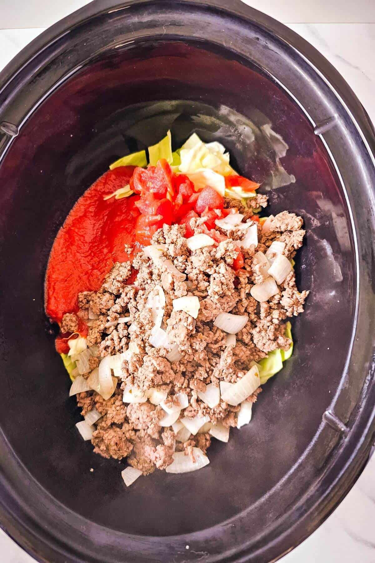 A slow cooker filled with ground meat, chopped onions, diced tomatoes, tomato sauce, and cabbage.