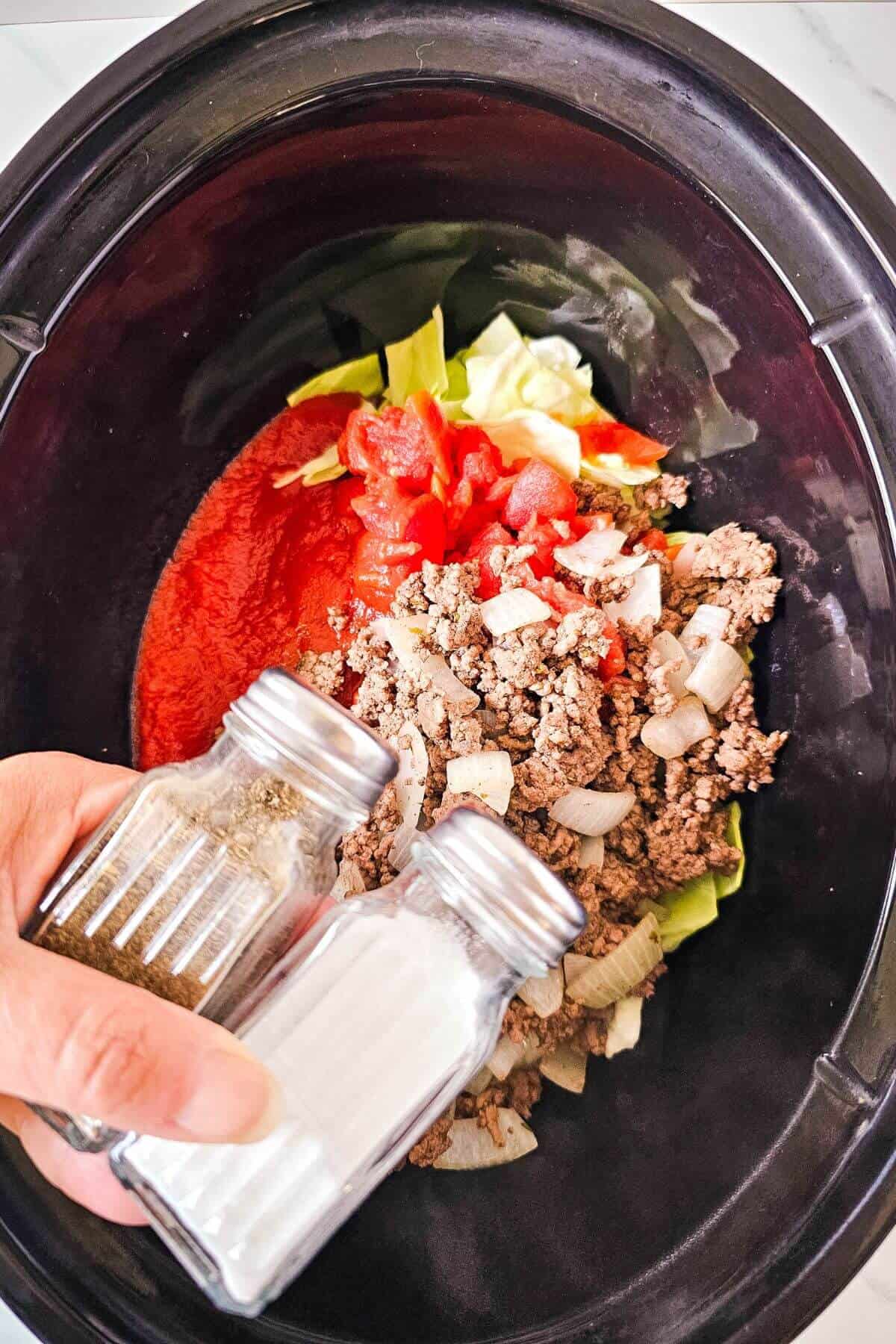 A hand holds salt and pepper shakers over a slow cooker containing ground beef, onions, cabbage, tomatoes, and sauce.