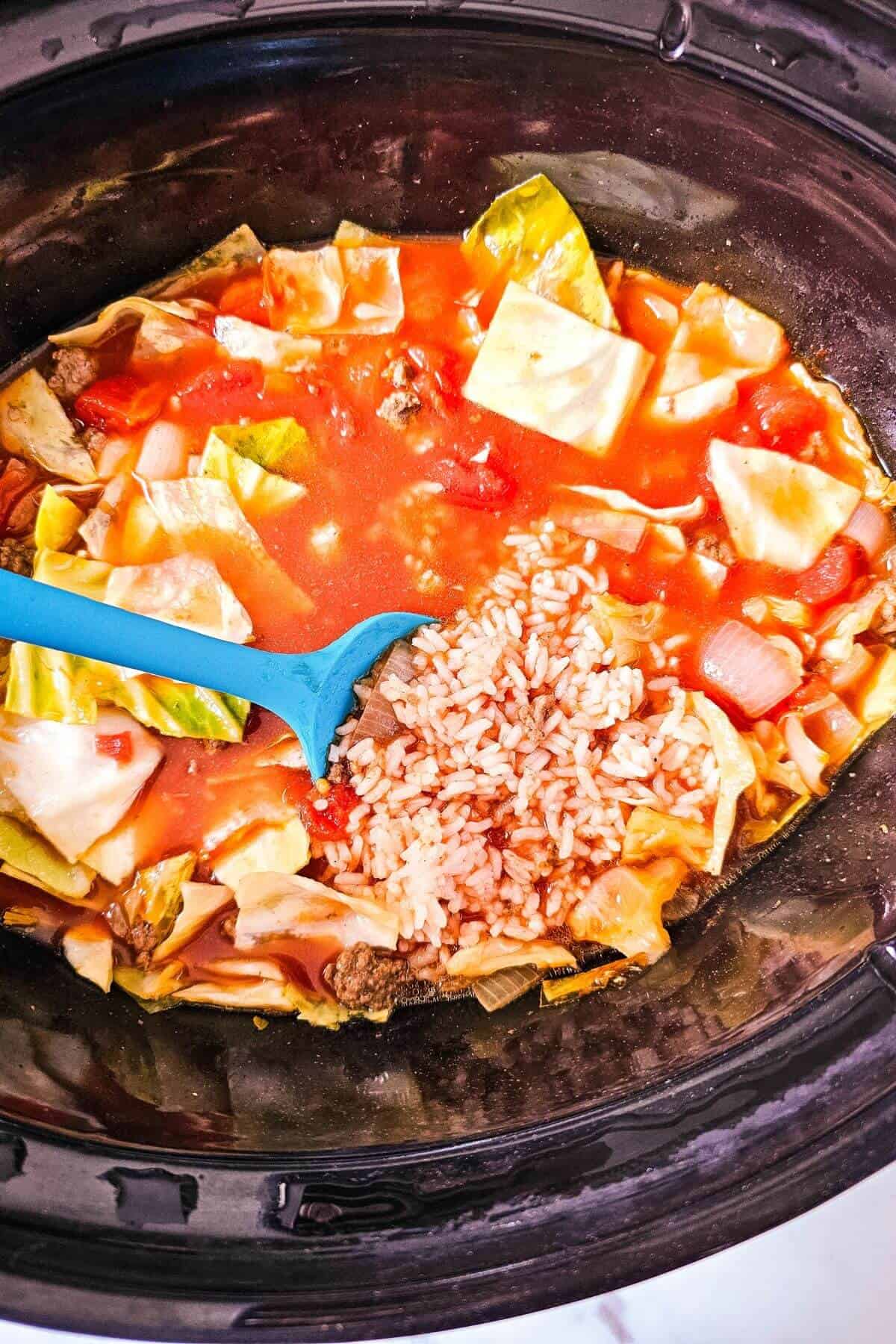 A blue spoon rests in a pot of cabbage soup containing chopped cabbage, rice, and a rich red broth.