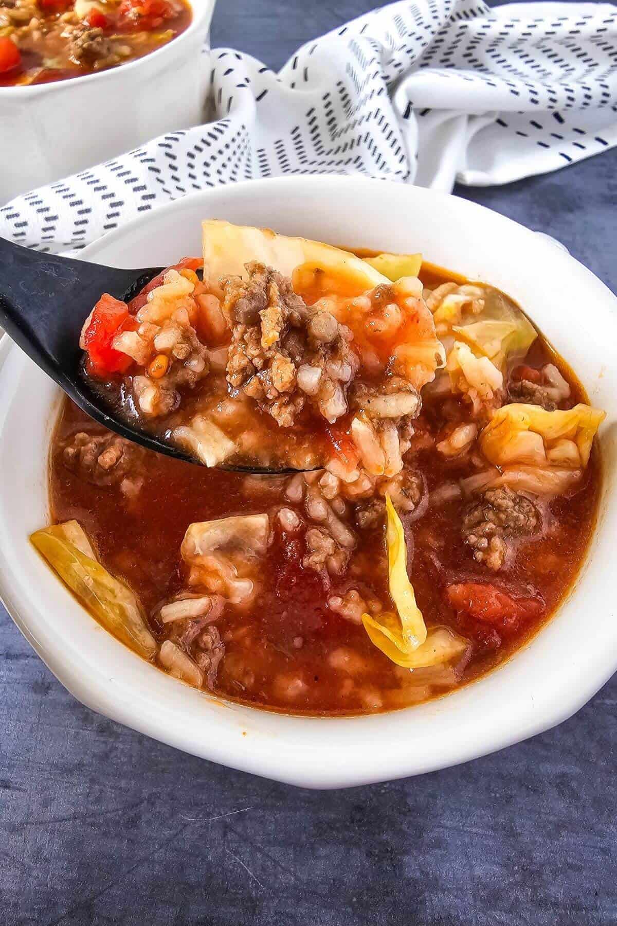 Close-up of a white bowl filled with cabbage soup, featuring ground meat, rice, and tomatoes. A black spoon holds a scoop of the soup above the bowl. A patterned cloth is in the background.