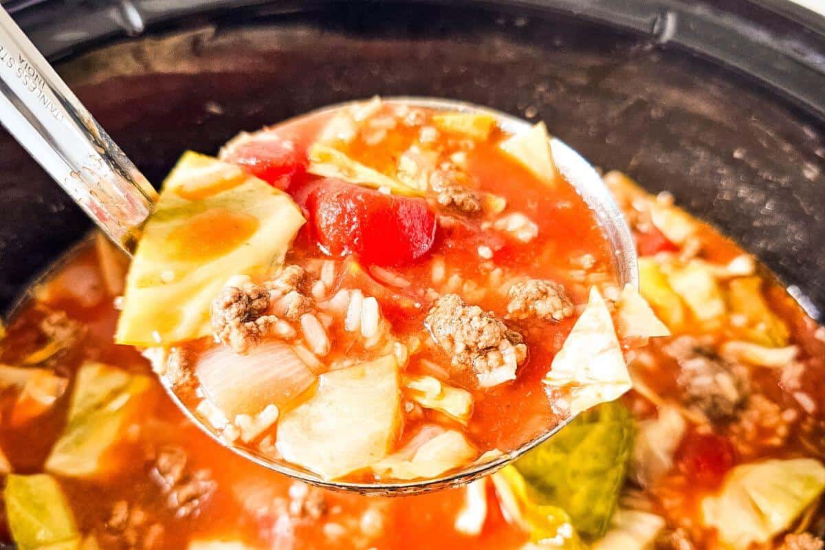 A ladle holding cabbage soup with ground beef, tomatoes, onions, and rice above a pot.