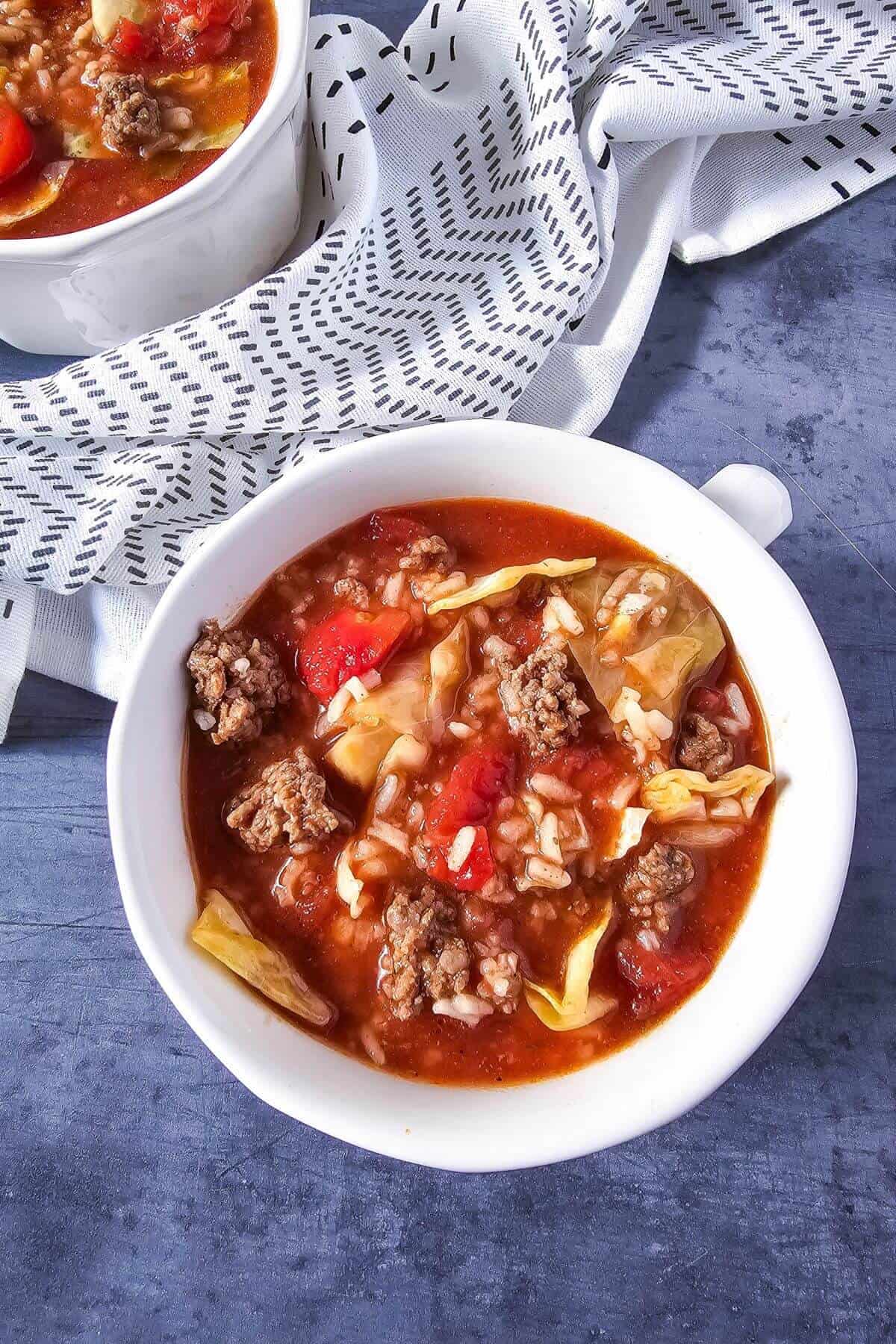 A bowl of soup with ground meat, cabbage, tomatoes, and rice, with a textured cloth in the background.
