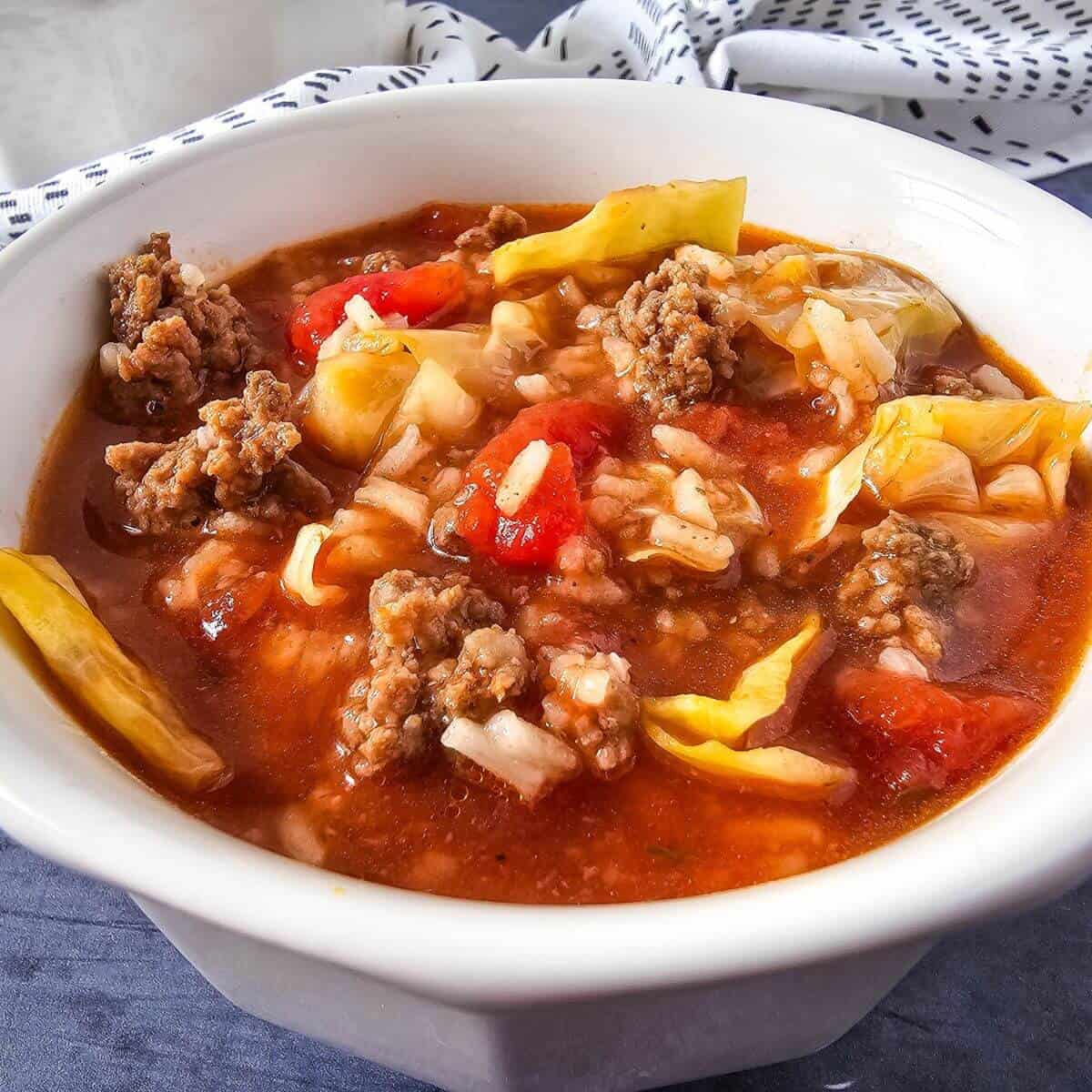 A bowl of soup with ground meat, cabbage, rice, and tomatoes in a red broth, set on a table with a patterned cloth.