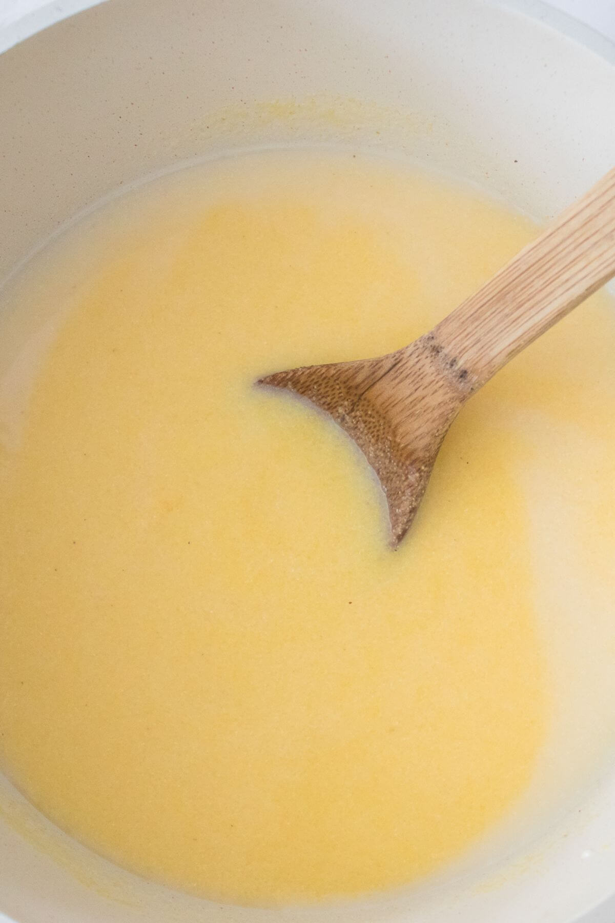A wooden spoon stirs creamy polenta in a white pot.