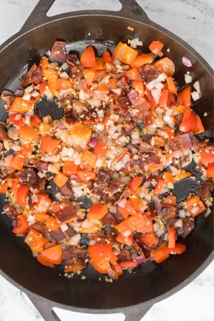 A skillet with diced bacon, red bell peppers, onions, and garlic sautéd together.