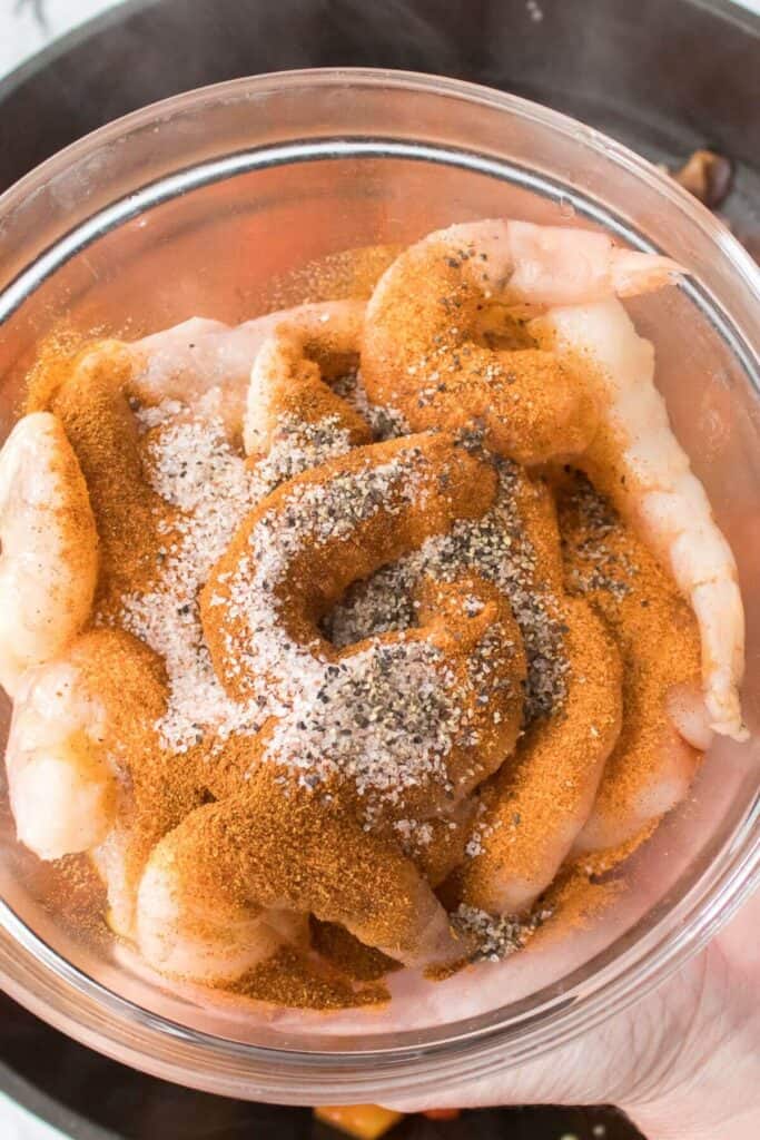 Raw shrimp covered with a mix of spices, including salt and pepper, in a glass bowl.