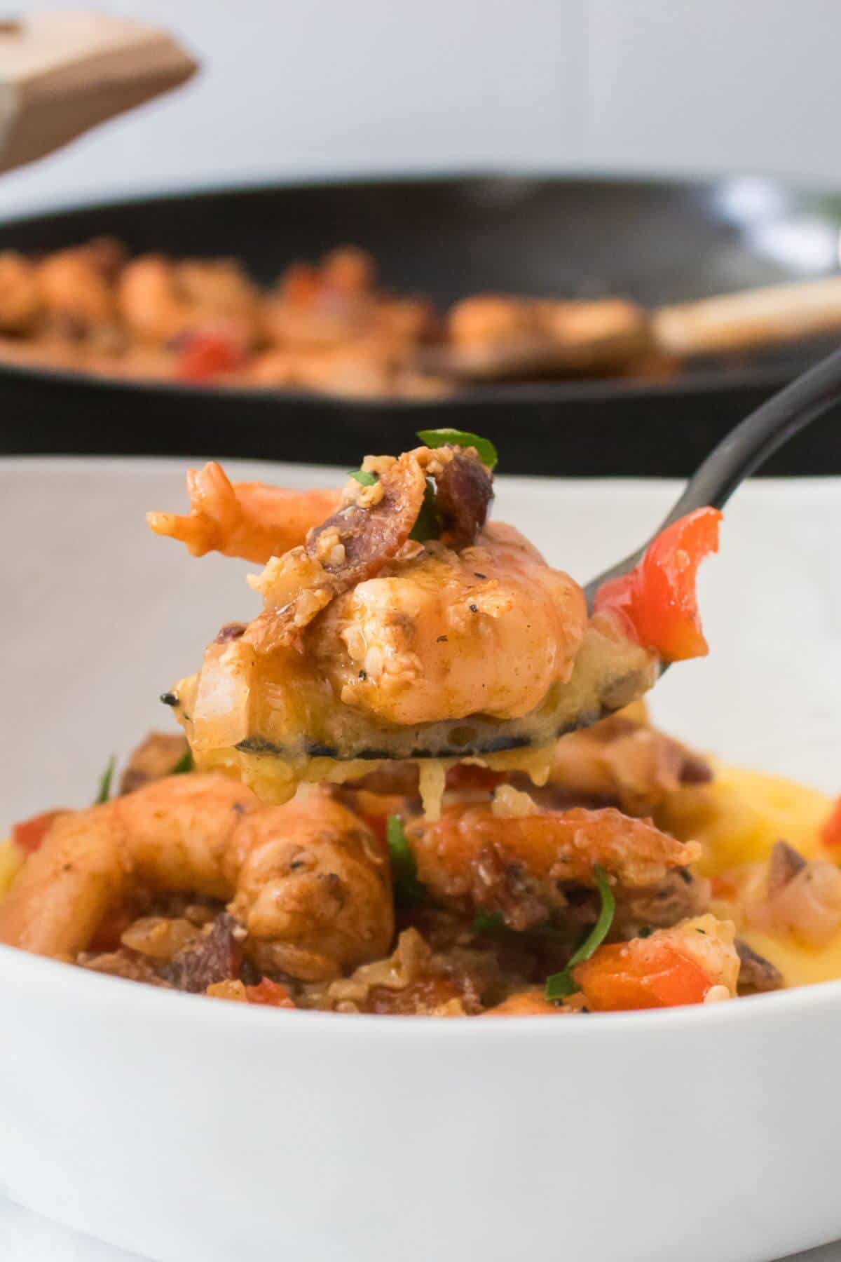 A spoonful of cooked shrimp with peppers and herbs is held over a white bowl, with a pan in the background.