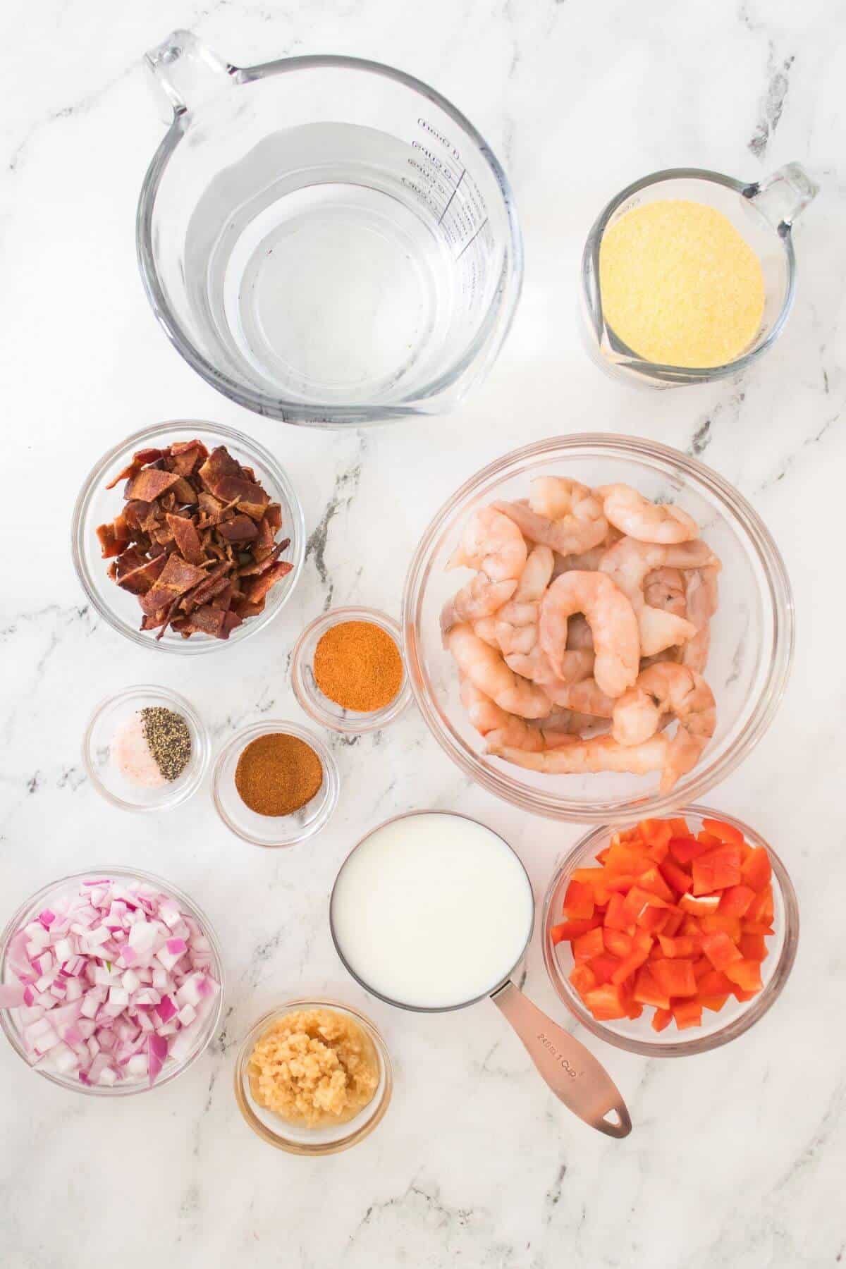 Image of cooking ingredients on a marble surface. Includes shrimp, diced red peppers, chopped onions, garlic, bacon, spices, cornmeal, water, and milk in various bowls and measuring cups.