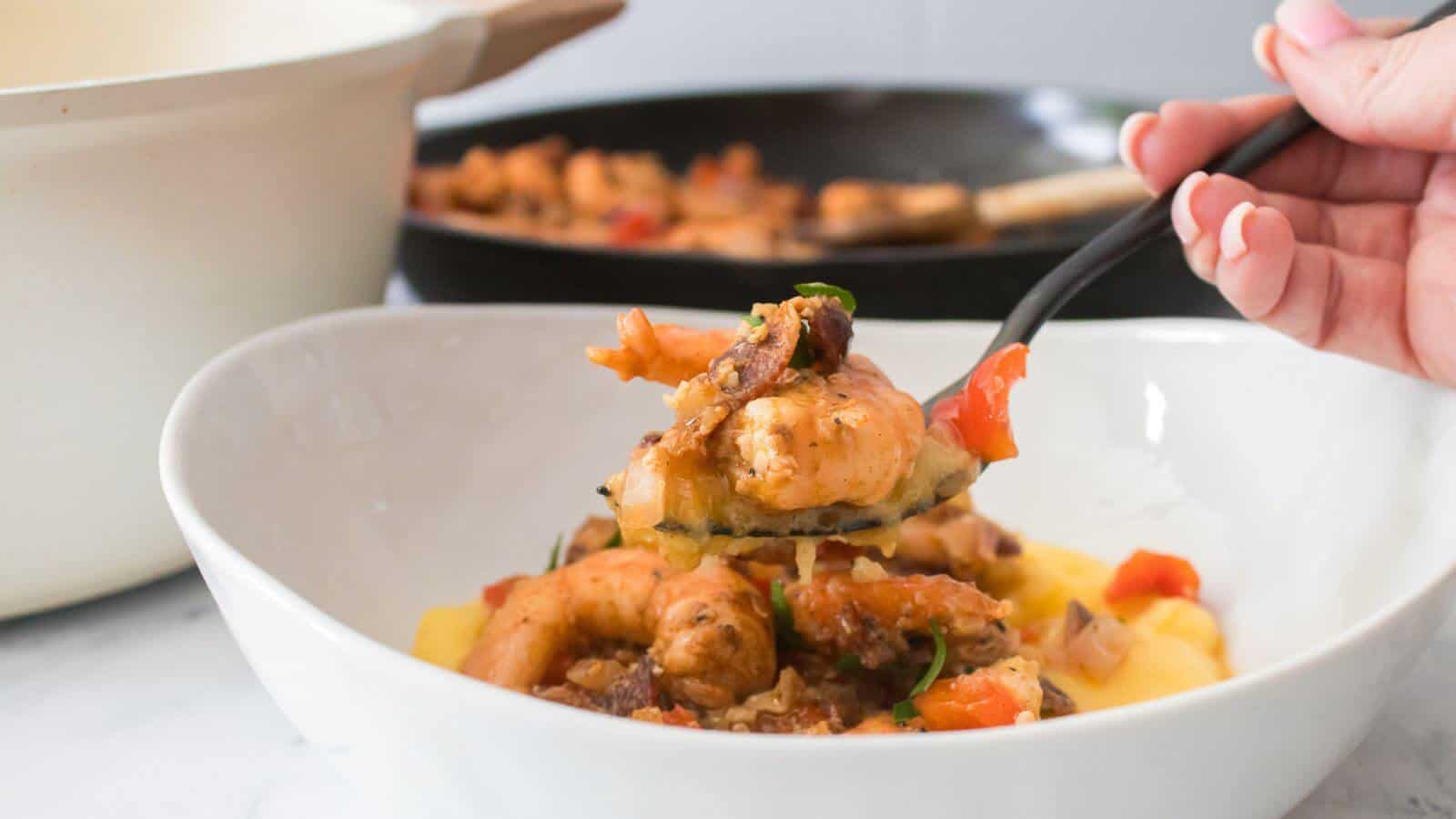A fork holds shrimp and vegetables over a bowl of creamy polenta, with more shrimp and vegetables in a skillet in the background.