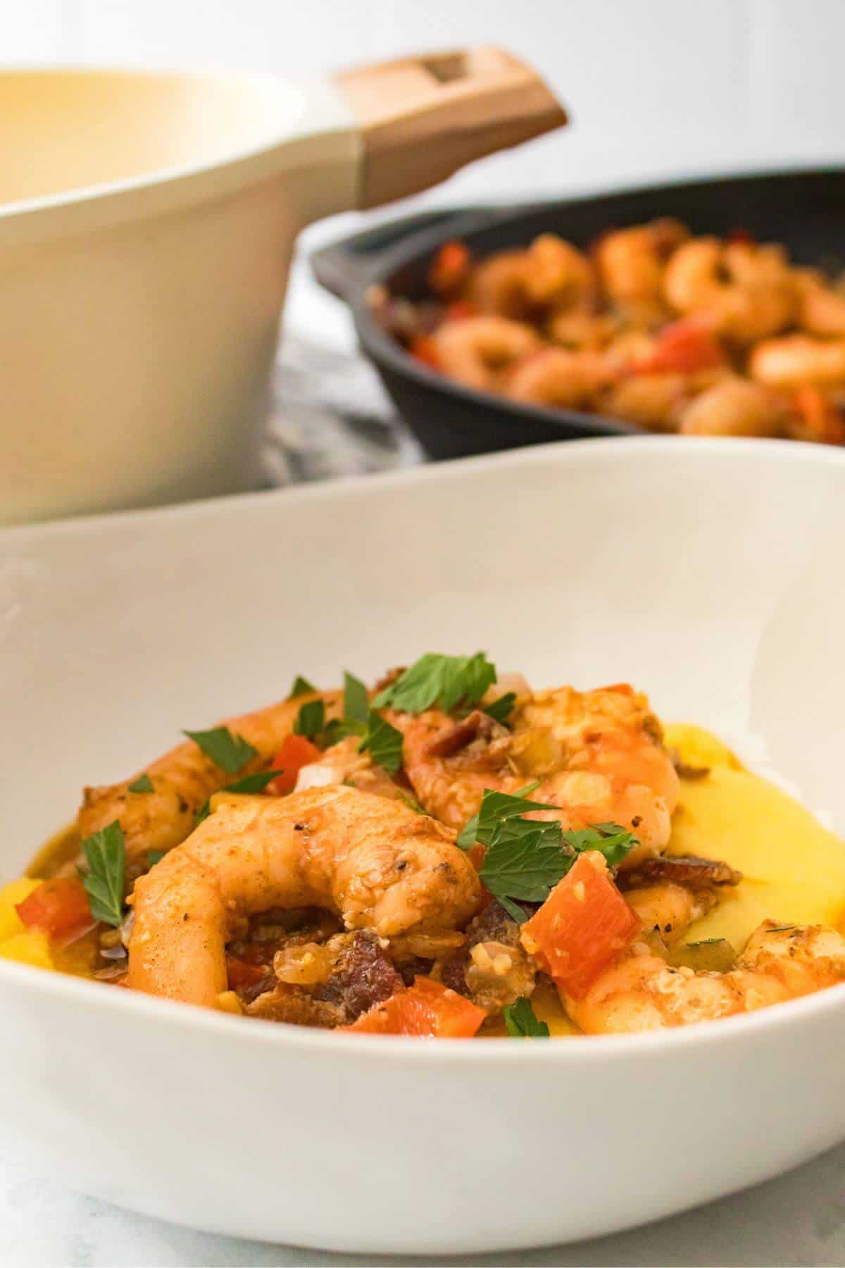 A bowl of shrimp with tomatoes and parsley on a bed of creamy polenta. A skillet with additional shrimp in the background.