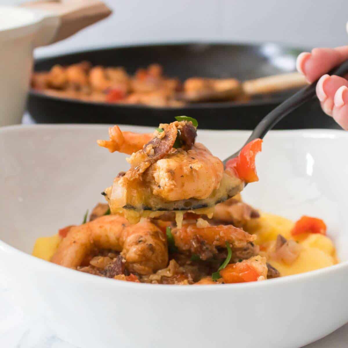 A spoonful of shrimp and vegetables is lifted from a bowl of creamy polenta. A pan of additional shrimp and vegetables is in the background.