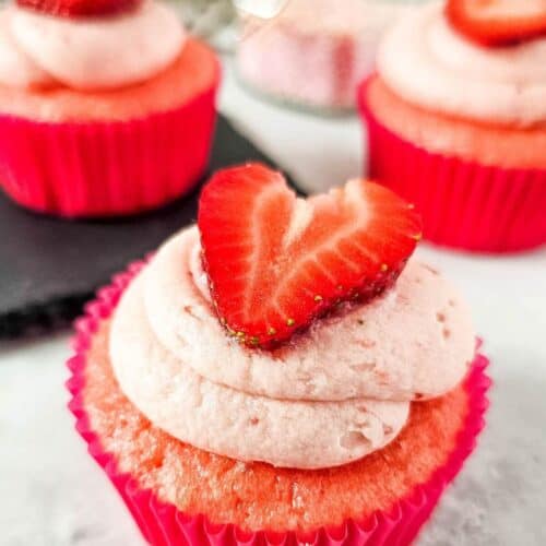 Strawberry cupcakes with pink frosting and heart-shaped strawberry slices on top, arranged on a white surface.