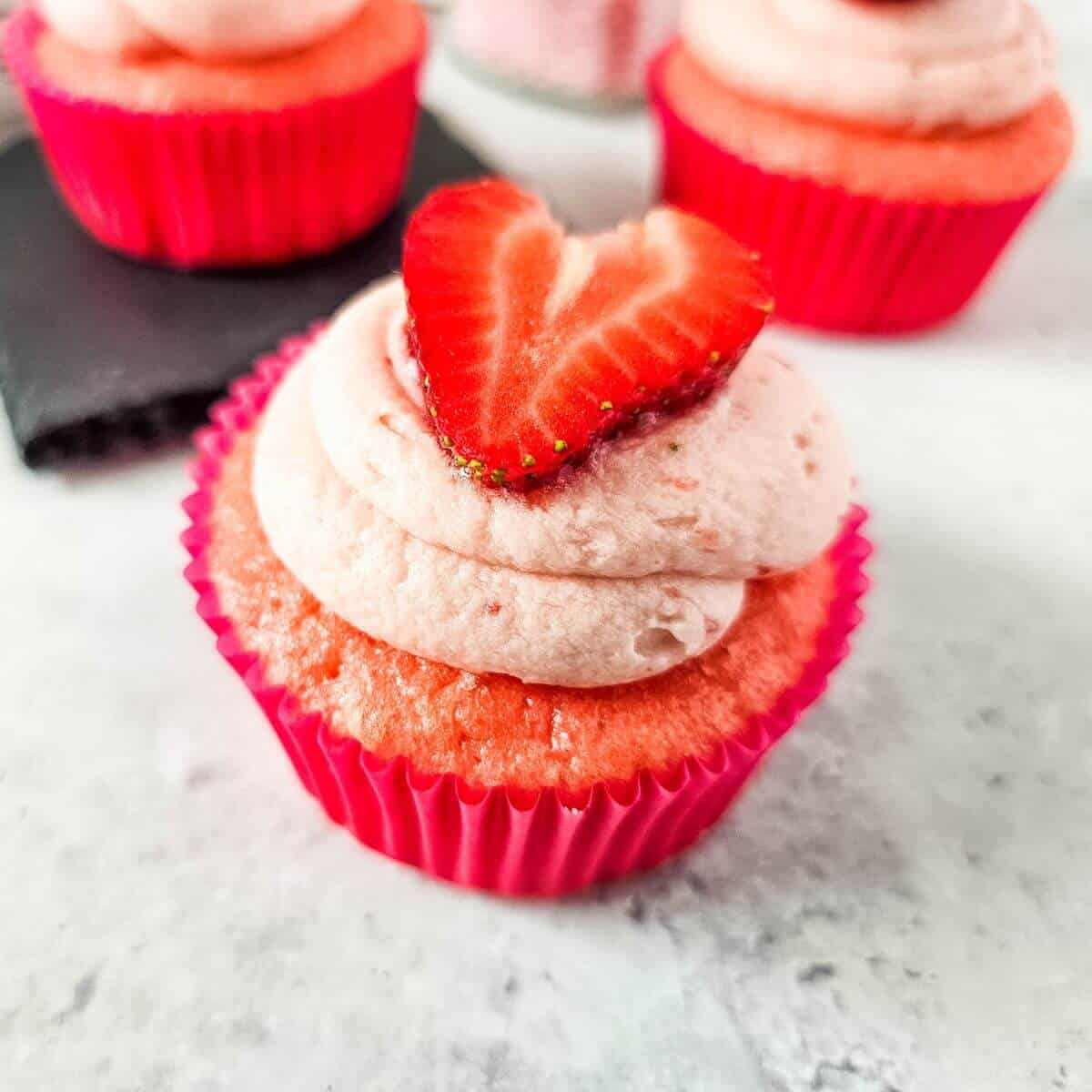 A pink cupcake with strawberry frosting and a heart-shaped strawberry slice on top, in a pink wrapper, set on a light surface.