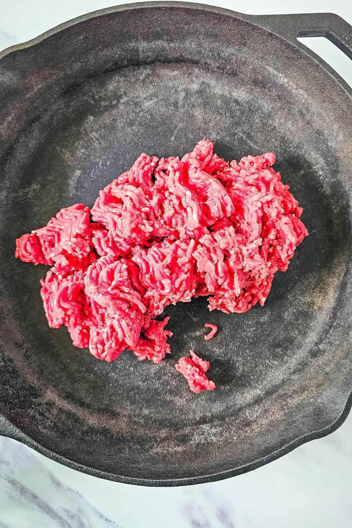 Raw ground beef in a black frying pan on a light-colored surface.