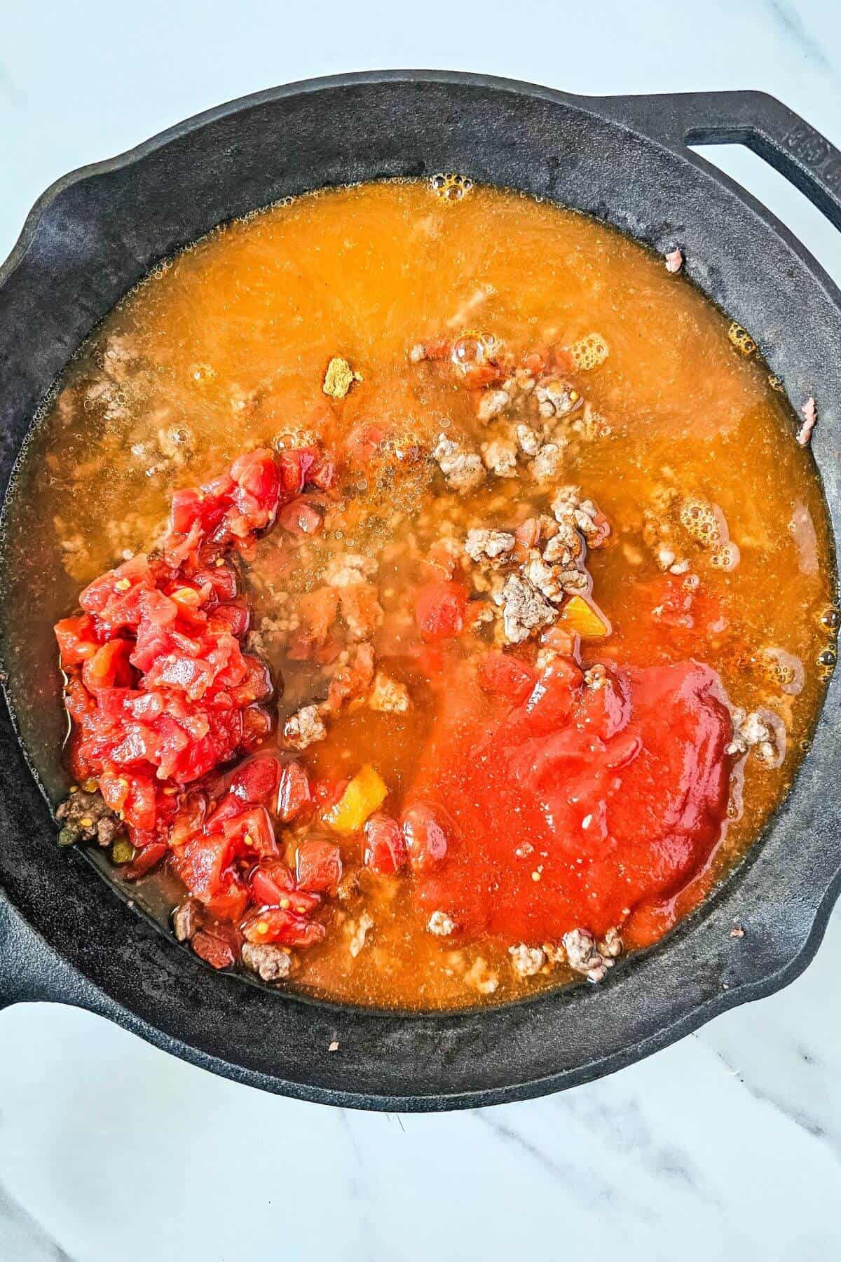 A cast iron skillet with ground beef, diced tomatoes, and tomato sauce cooking together.