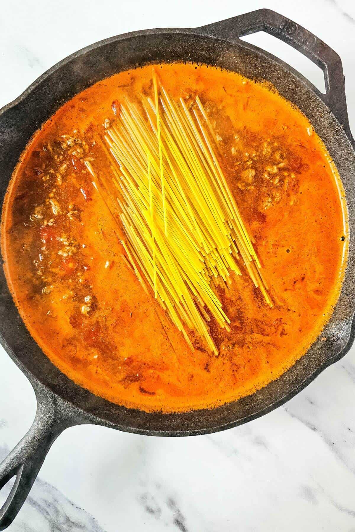 A cast iron skillet with uncooked spaghetti and a tomato-based sauce with ground meat, simmering on a stove.