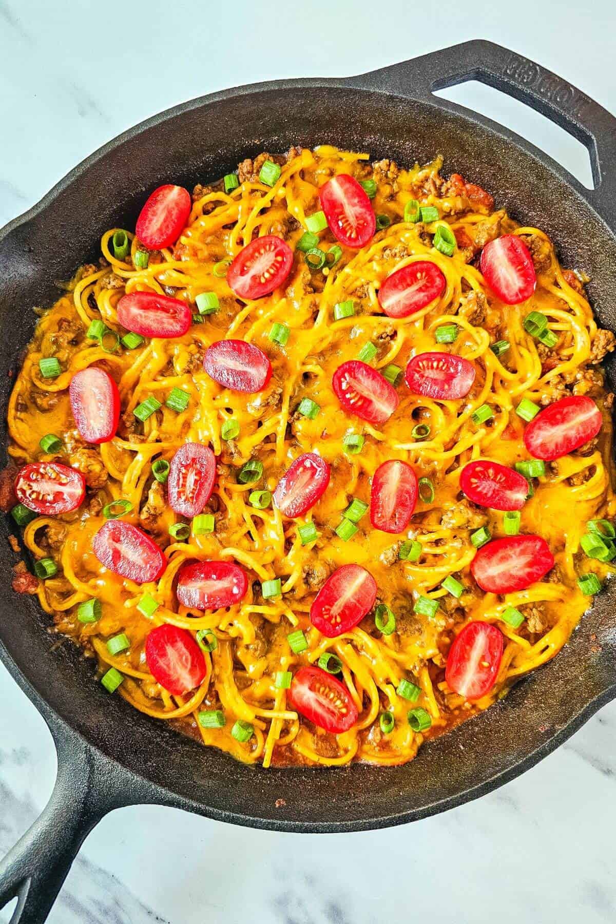 Cast iron skillet with spaghetti topped with melted cheese, sliced cherry tomatoes, and chopped green onions.