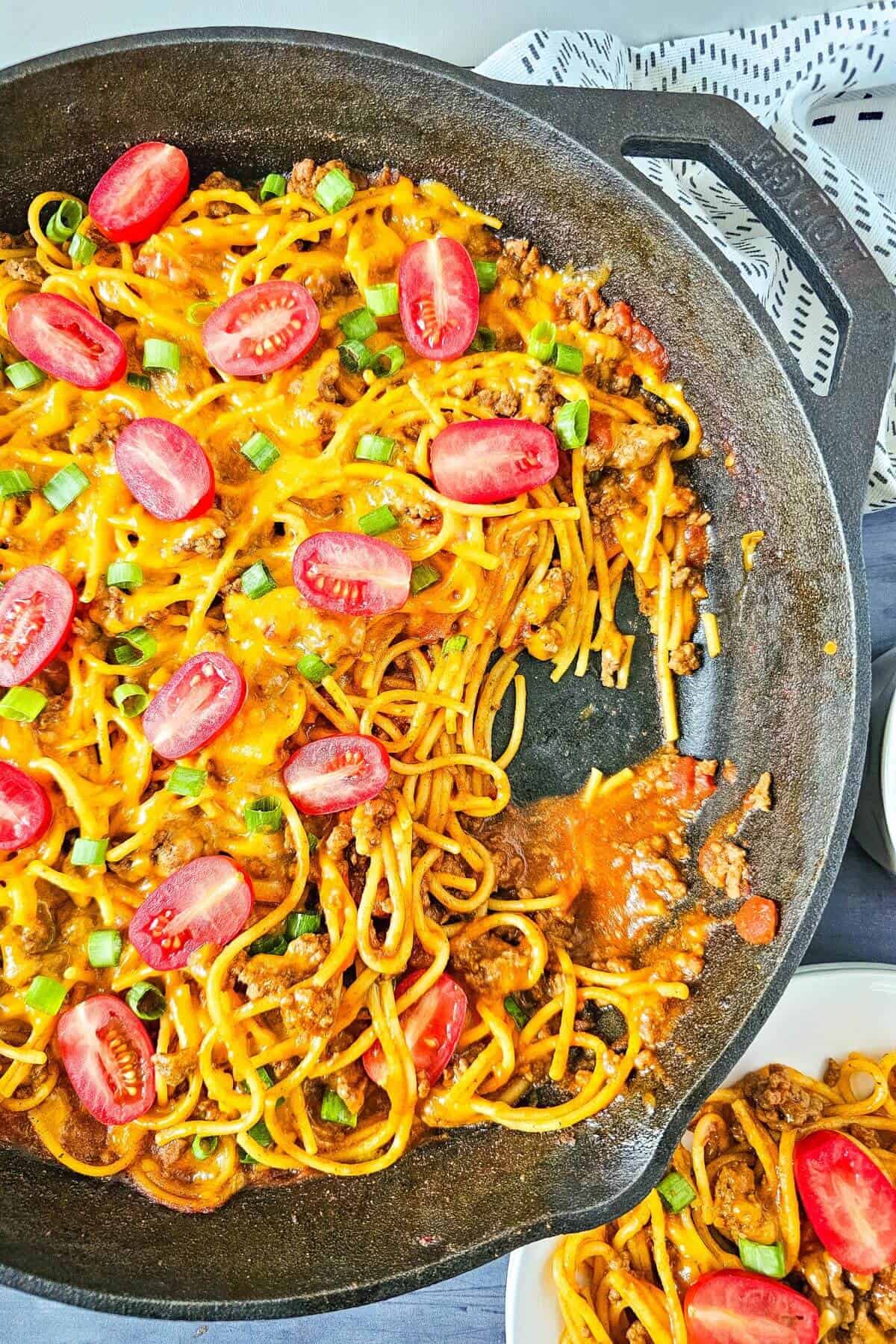 A cast iron skillet filled with cheesy baked spaghetti topped with sliced cherry tomatoes and chopped green onions.