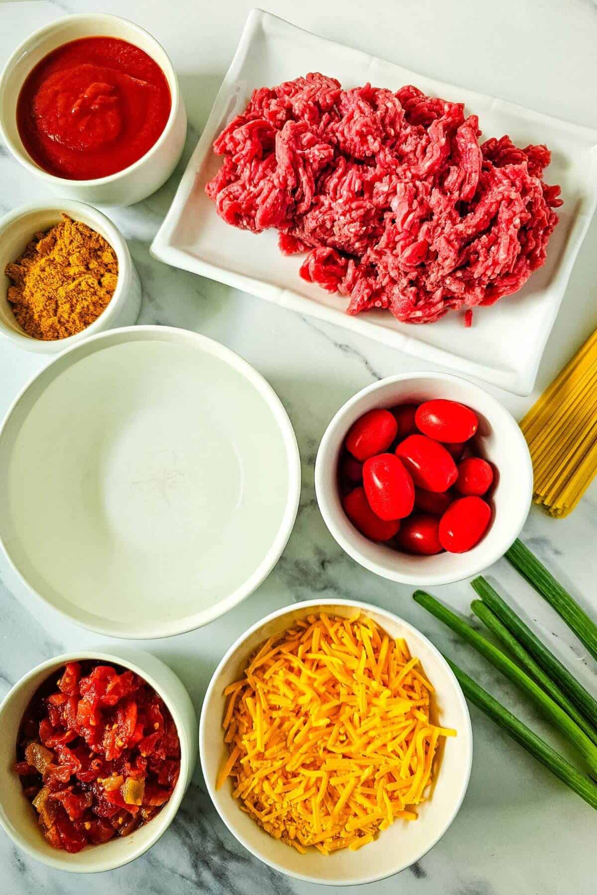 Ingredients on a countertop include ground meat, cherry tomatoes, shredded cheese, diced tomatoes, taco seasoning, tomato sauce, green onions, water, and uncooked spaghetti.