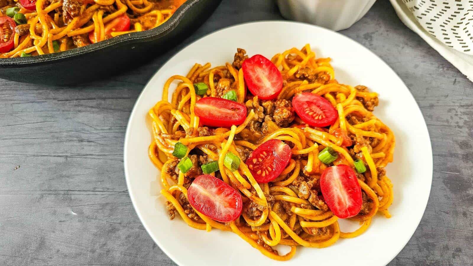 A plate of spaghetti with ground meat, halved cherry tomatoes, and chopped green onions. A black skillet containing more of the dish is partially visible in the background.