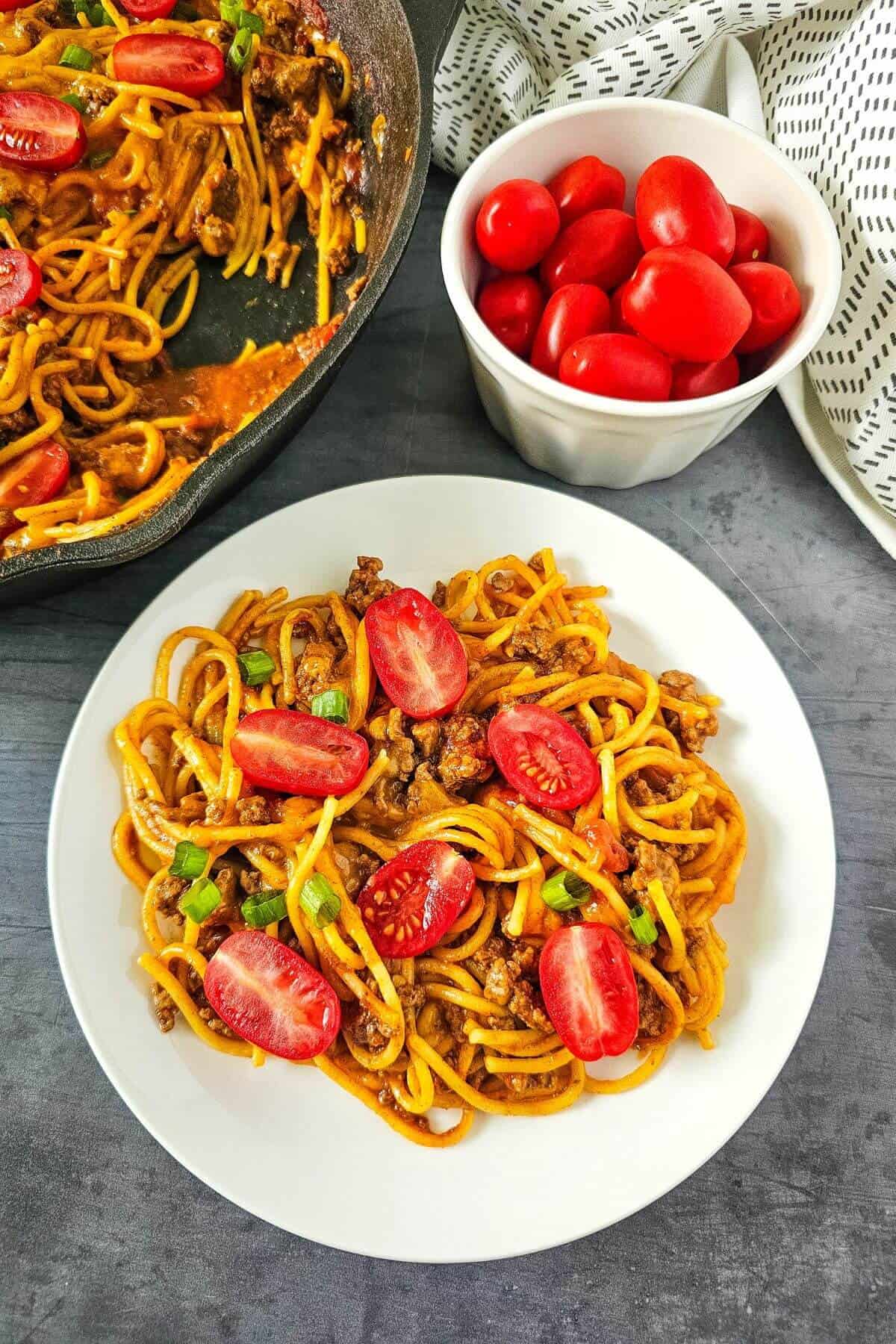 Plate of spaghetti with ground meat and cherry tomatoes beside a bowl of fresh cherry tomatoes and a pan on a gray surface.