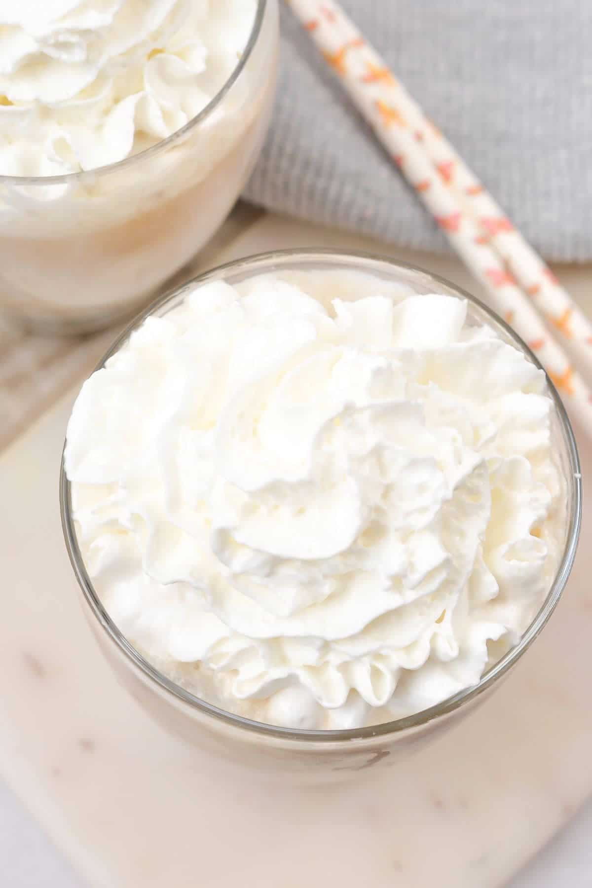 Glass of whipped cream beside a chilled iced white chocolate mocha with paper straws in the background on a light surface.