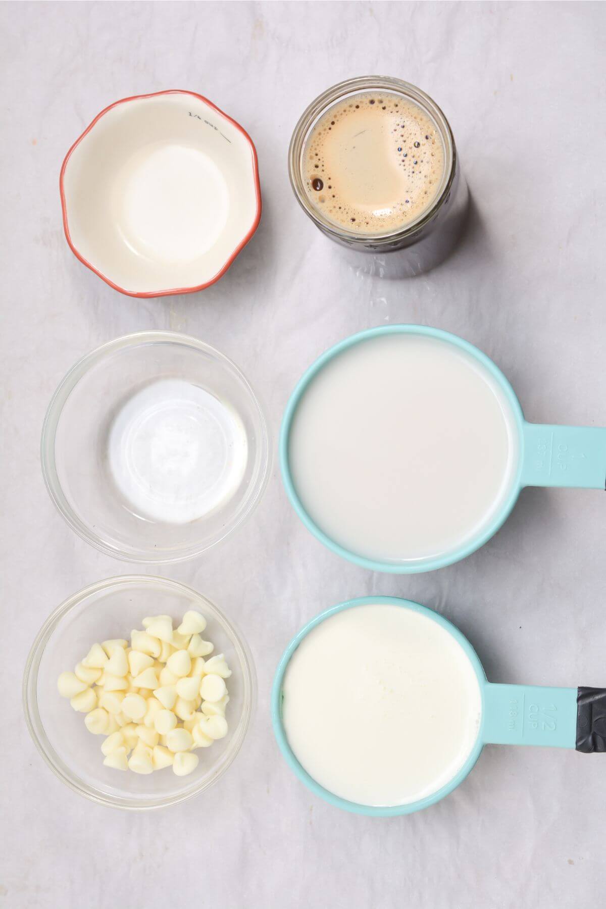 Ingredients arranged on a surface: a bowl of white chocolate chips, two measuring cups of milk, a glass of iced coffee, perfect for making that refreshing iced white chocolate mocha. Nearby are a small empty bowl and a small bowl with cream.