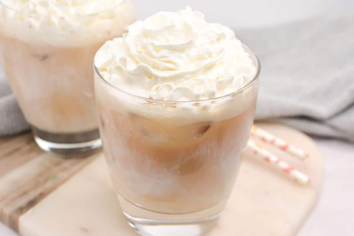 Two glasses of iced white chocolate mocha, each topped with whipped cream, sit elegantly on a wooden cutting board, with striped straws beside them.