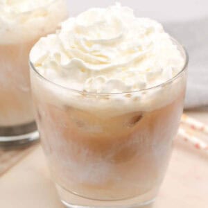 Glass of iced white chocolate mocha topped with whipped cream on a light surface, with another drink and paper straws in the background.