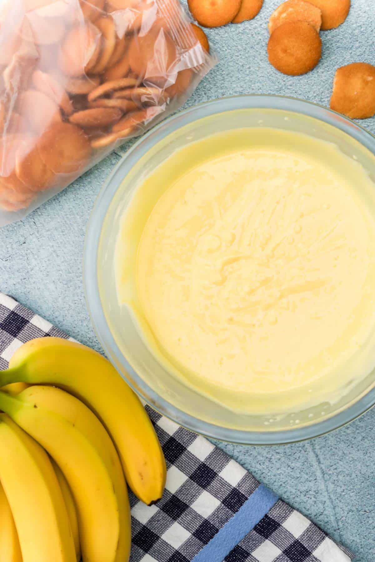 A bowl of creamy banana pudding is surrounded by a bunch of bananas, a bag of vanilla wafers, and some scattered cookies on a checkered cloth.