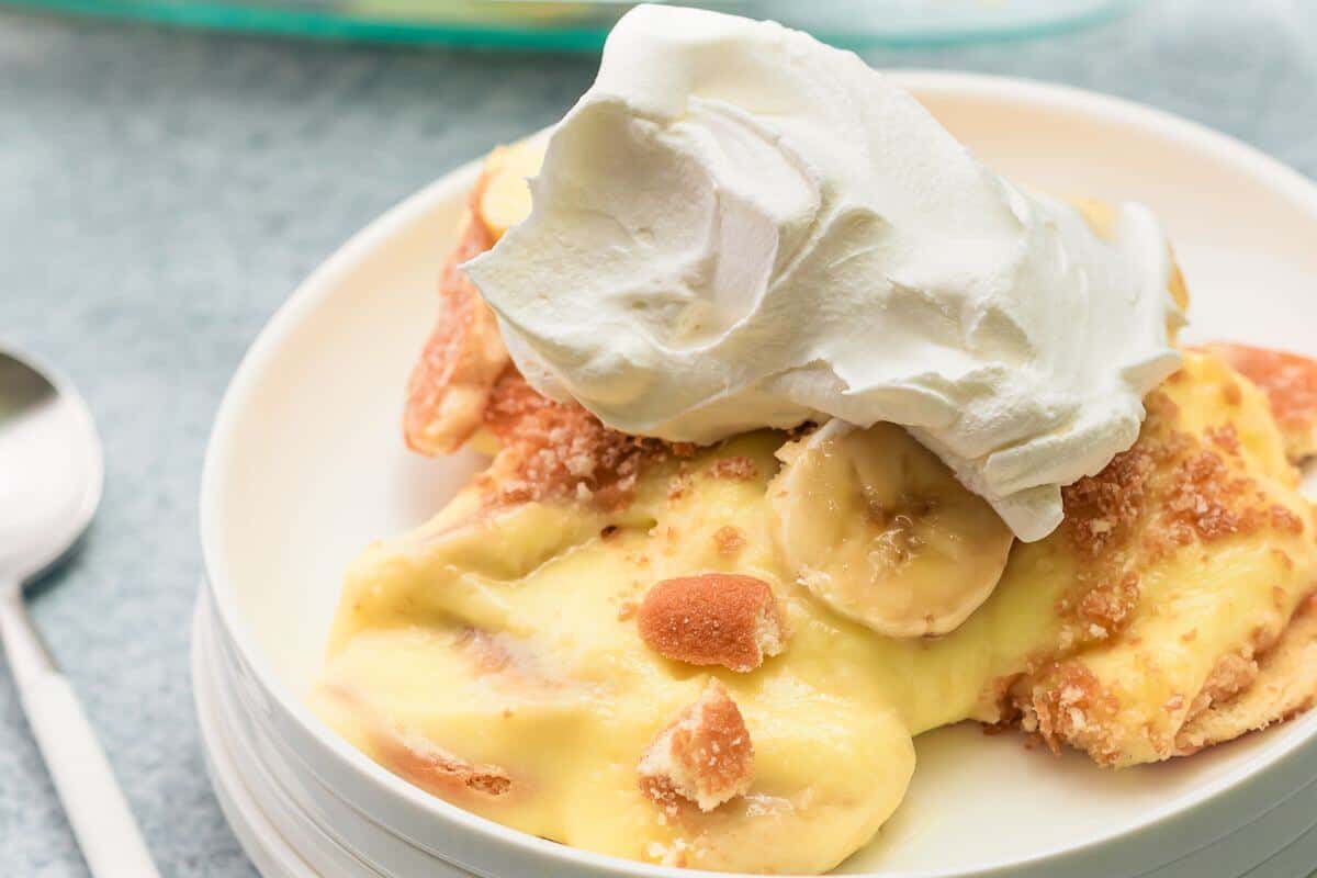 A serving of banana pudding topped with whipped cream on a white plate, with a spoon nearby.