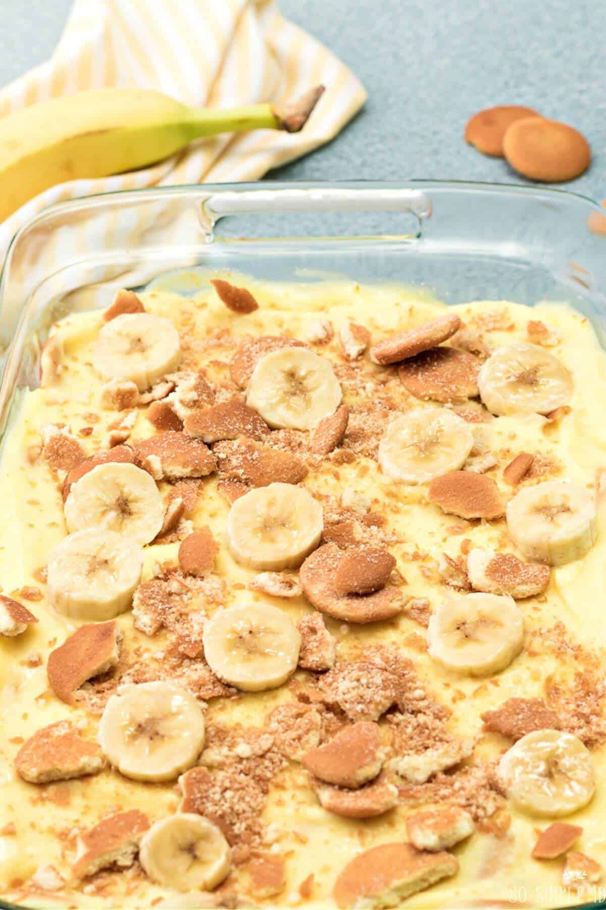 Banana pudding in a glass dish topped with banana slices and crumbled cookies, with a whole banana and a striped cloth in the background.