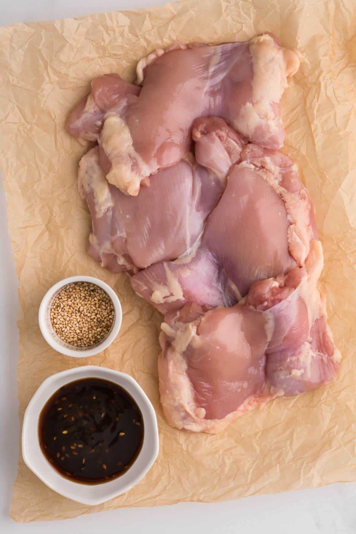 Raw chicken thighs on parchment paper with small bowls of sesame seeds and a dark sauce.