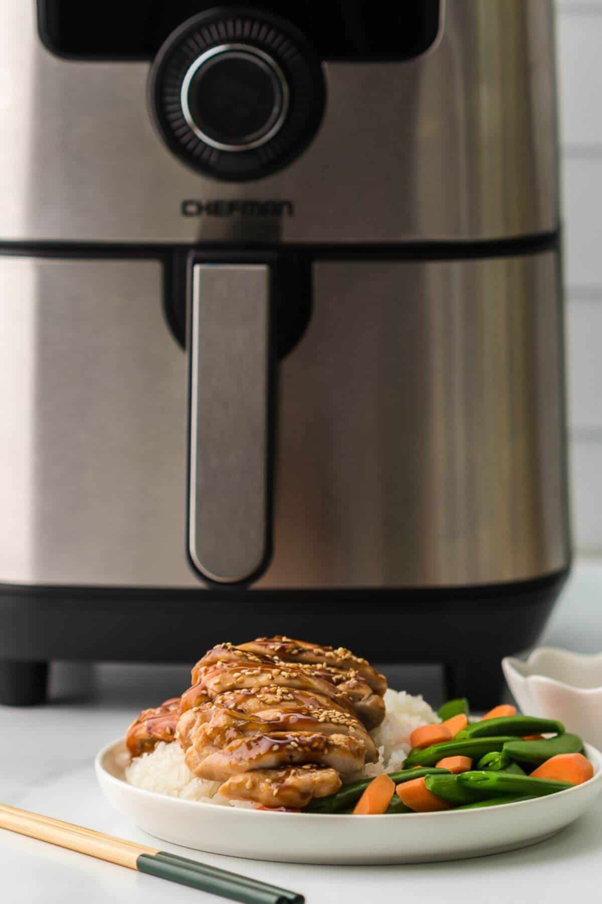 A plate with teriyaki chicken, rice, and vegetables is placed in front of a modern air fryer.