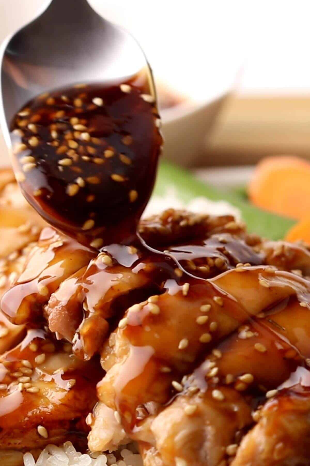A spoon drizzles a glossy, dark sauce with sesame seeds over pieces of cooked chicken.