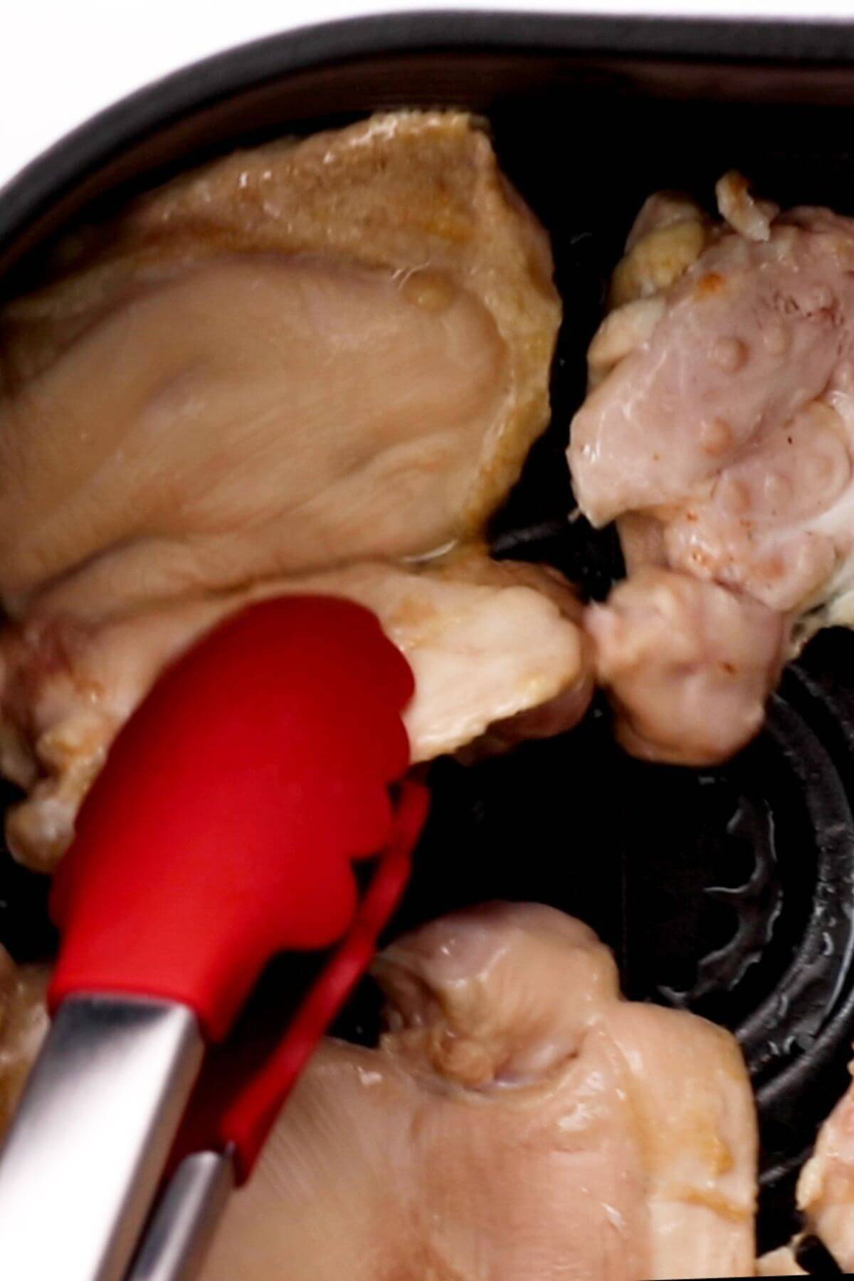 Red tongs holding a piece of raw chicken in a pan with other chicken pieces.