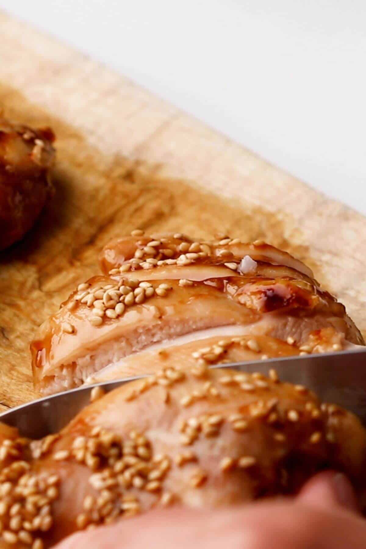 A knife slices through a piece of cooked chicken breast topped with sesame seeds on a wooden cutting board.