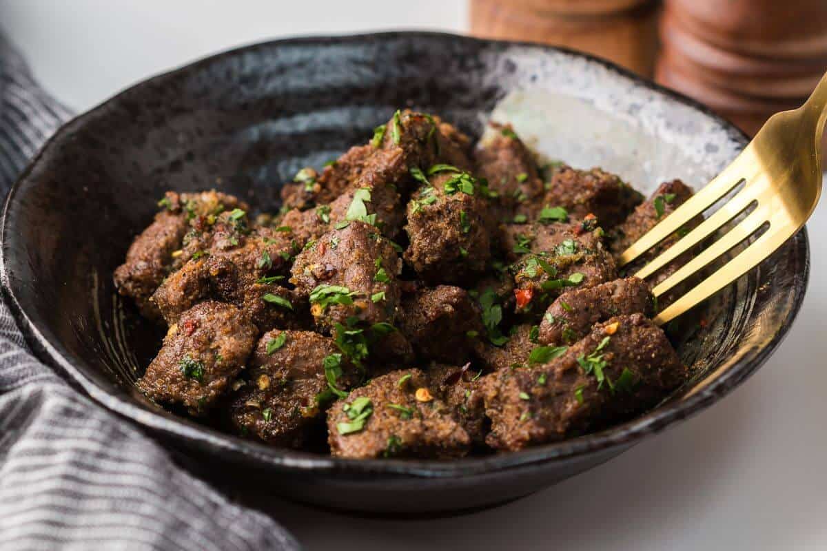 A bowl of seasoned meat cubes garnished with herbs, with a gold fork resting on the side.