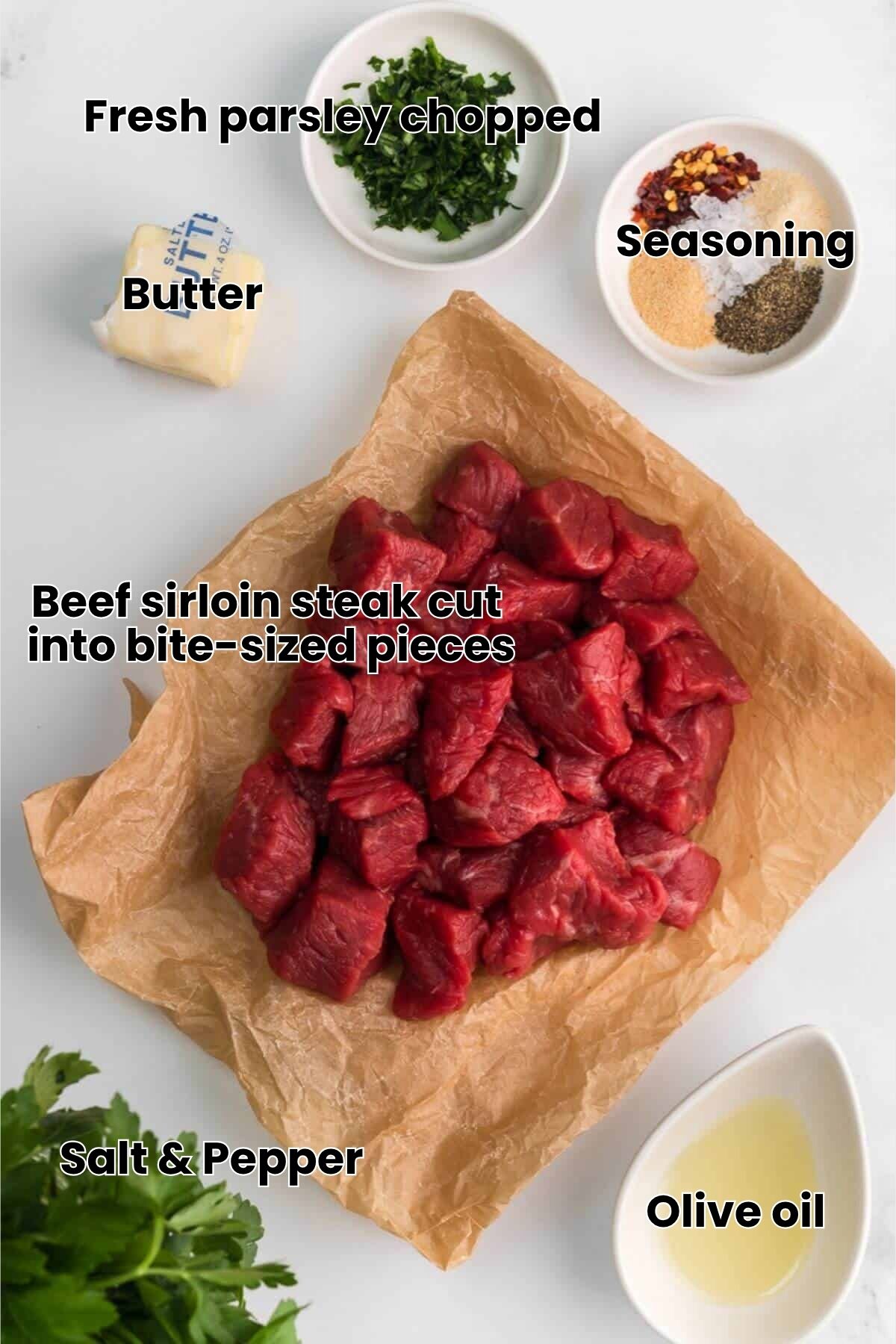 Raw beef cubes on parchment paper, surrounded by butter, chopped herbs, spices in small bowls, and a liquid in a white dish on a white background.